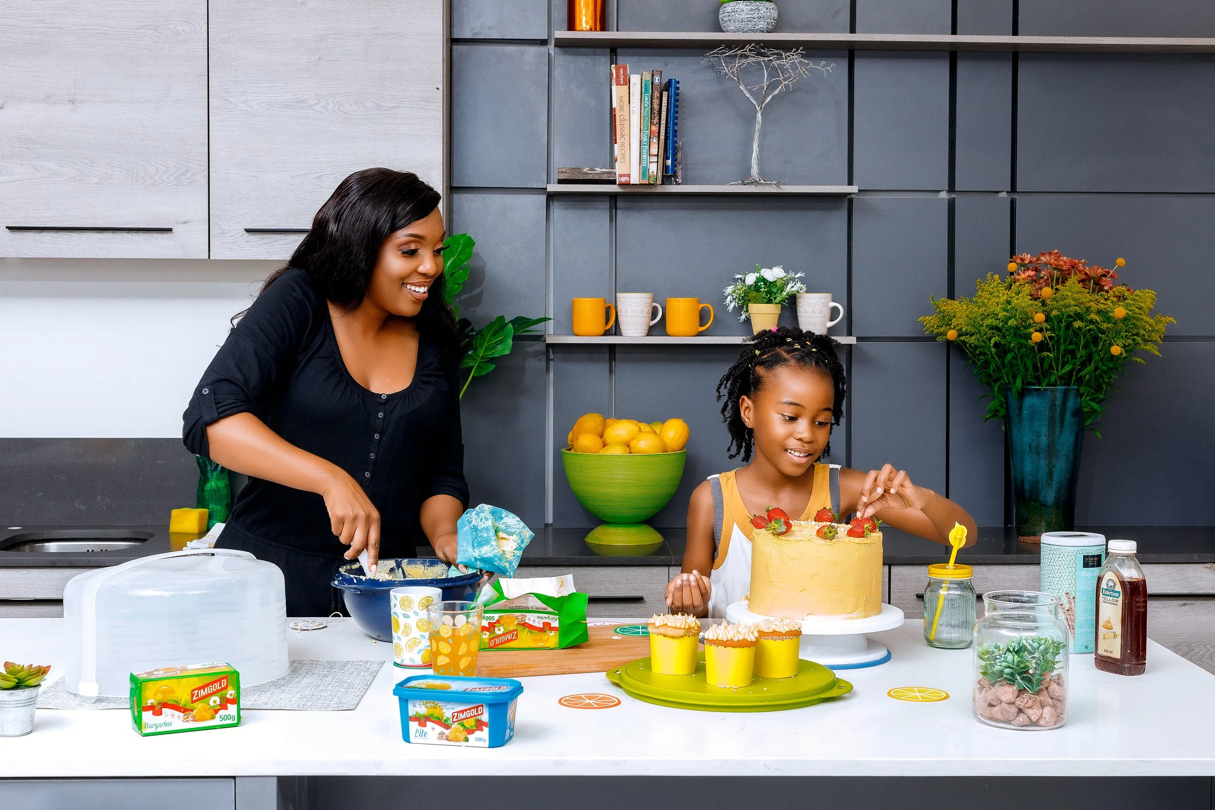 A woman and young girl baking and decorating a cake in a modern kitchen with various baked goods, ingredients, and kitchen items on the countertop.