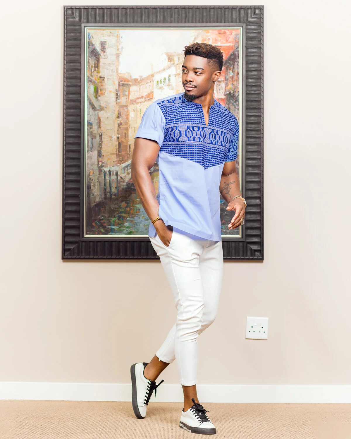 A young man dressed in a blue patterned shirt, white pants, and sneakers standing in front of a framed painting of a cityscape.