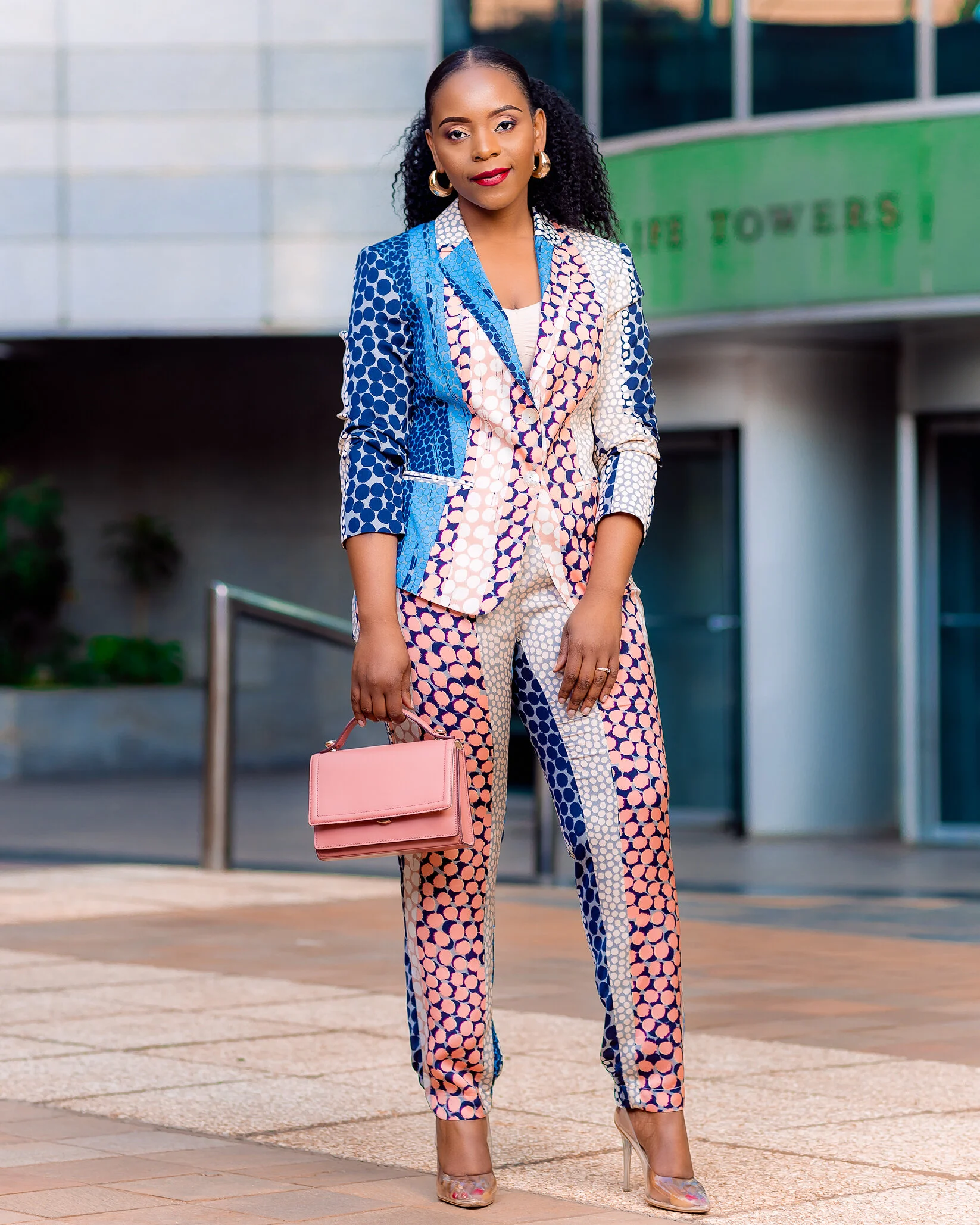 A woman wearing a colorful patterned suit consisting of a blazer and pants, holding a pink purse, standing outdoors in front of a modern building with glass windows and a green sign, with high-heeled shoes.