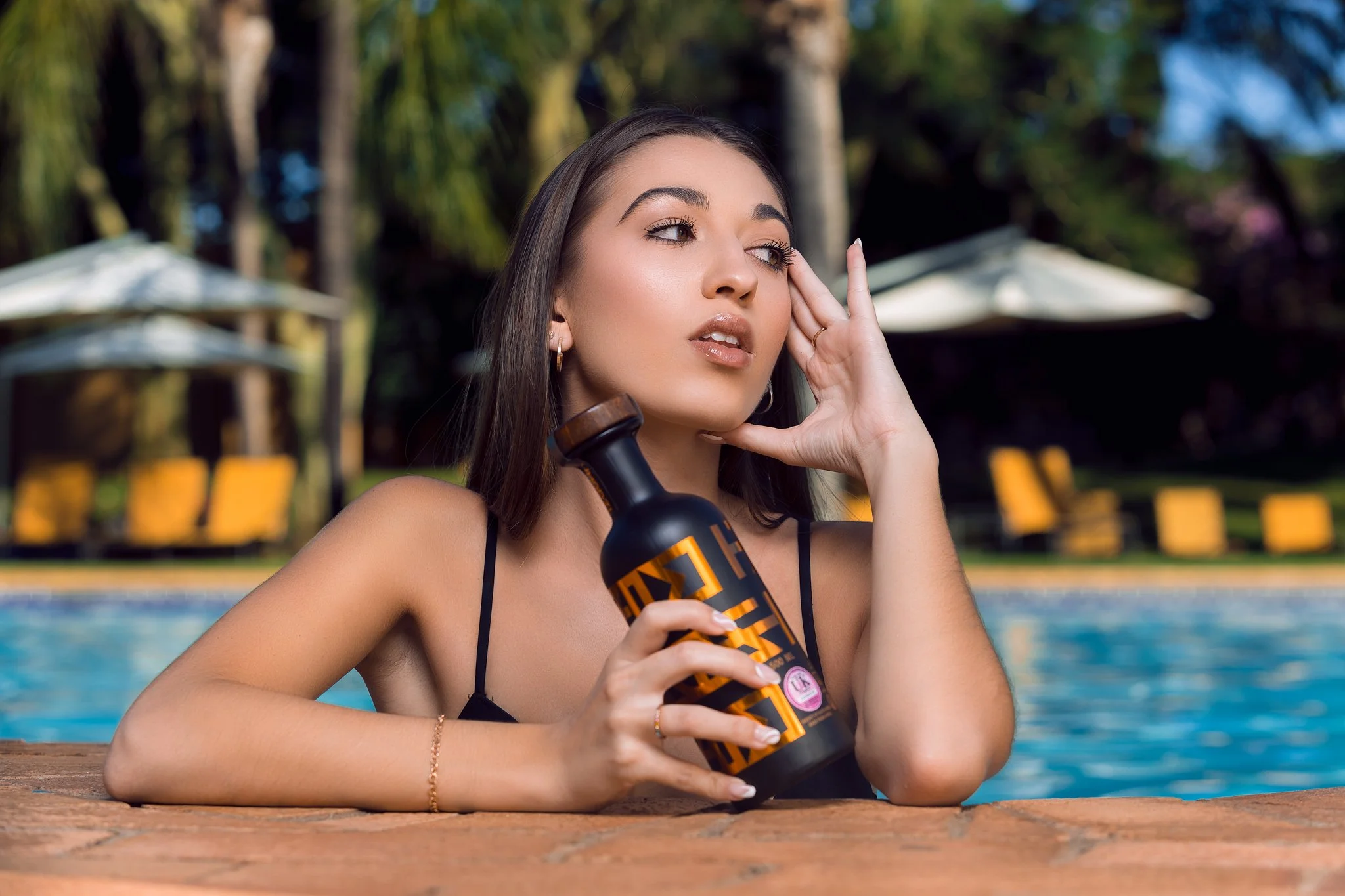 Young woman relaxing by a swimming pool holding a bottle of Hydroxycut dietary supplement, with poolside chairs and umbrellas in the background.