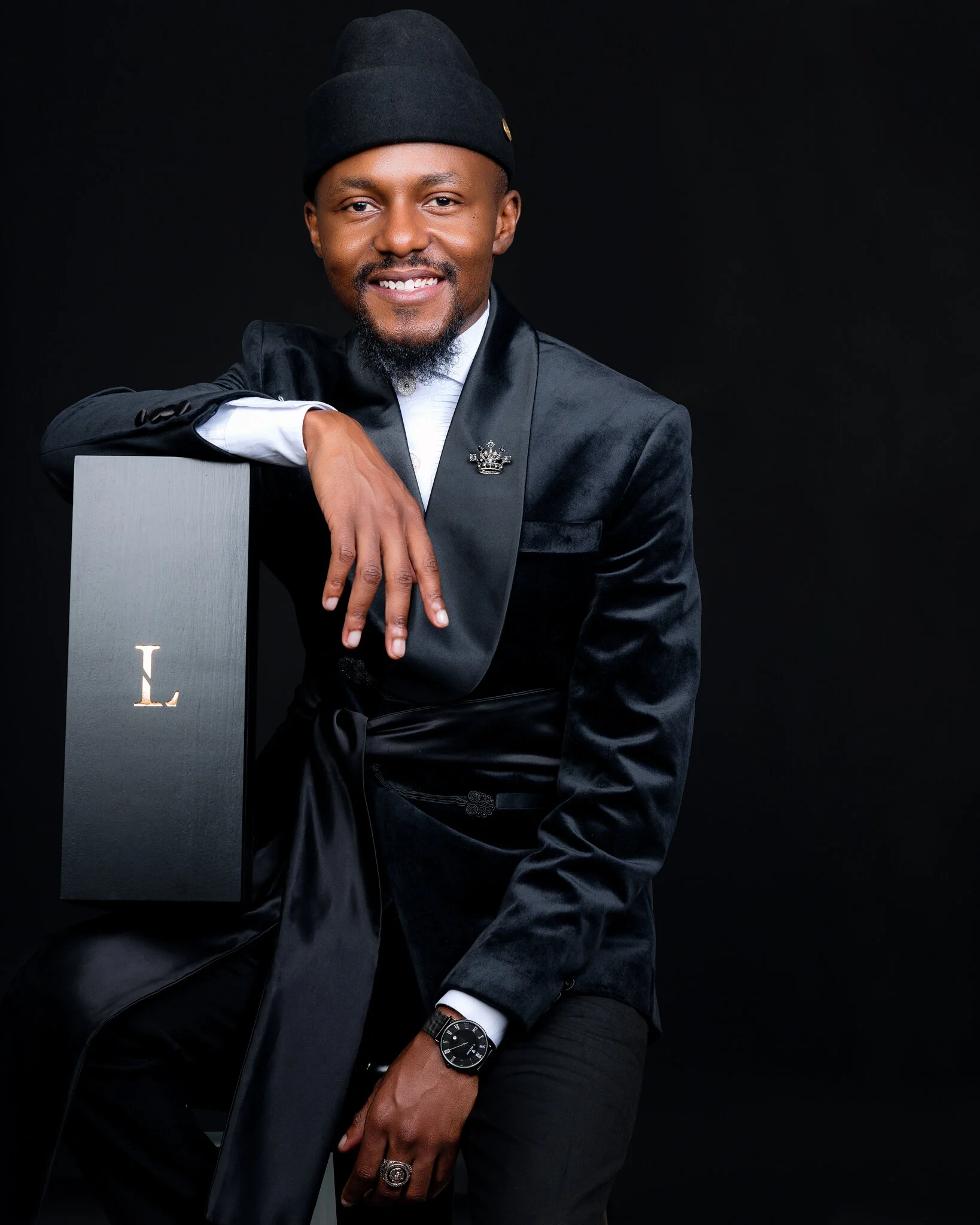 A man in a black beanie and black suit sitting against a black background, smiling, with his left elbow on a gray box with a gold 'L' on it, wearing a wristwatch and a ring.