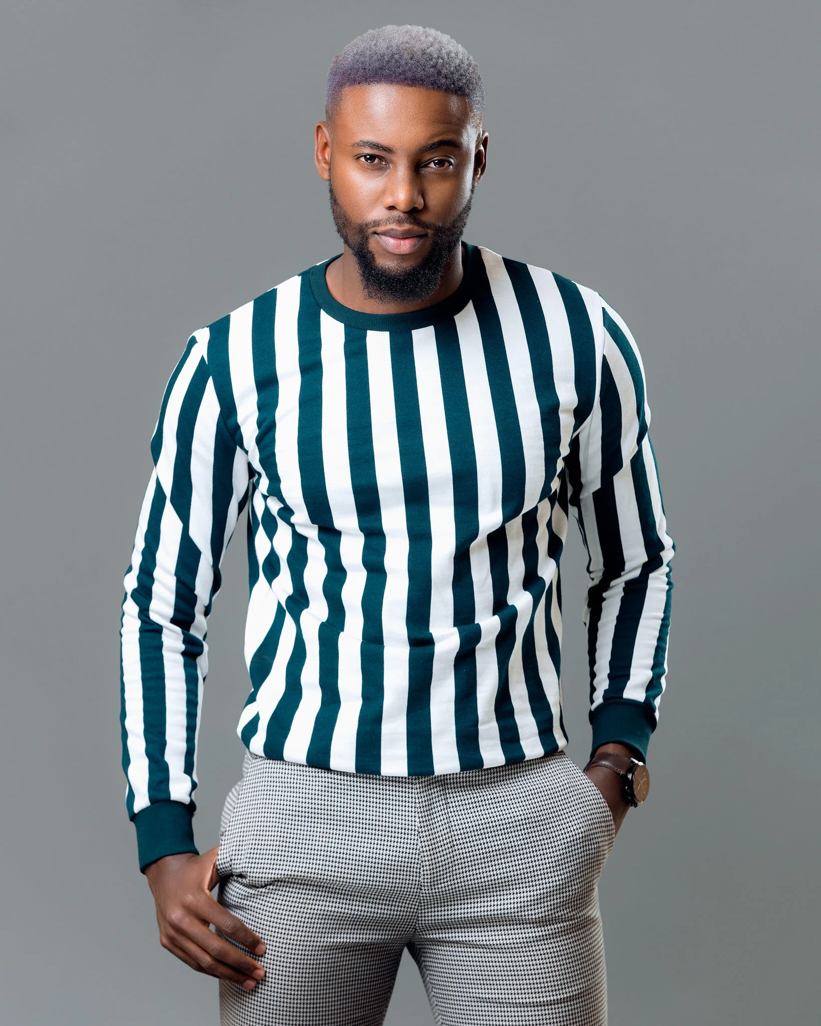 A man with a beard and dyed silver hair wearing a black and white vertically striped long sleeve shirt and checkered pants, standing against a grey background.