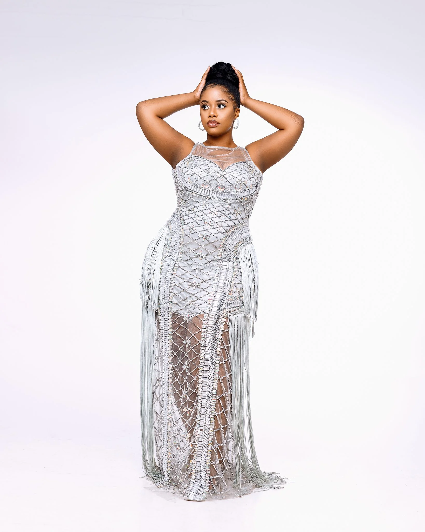 A woman in a silver, intricately beaded dress with fringe, posing with her arms raised and hands on her head against a plain white background.