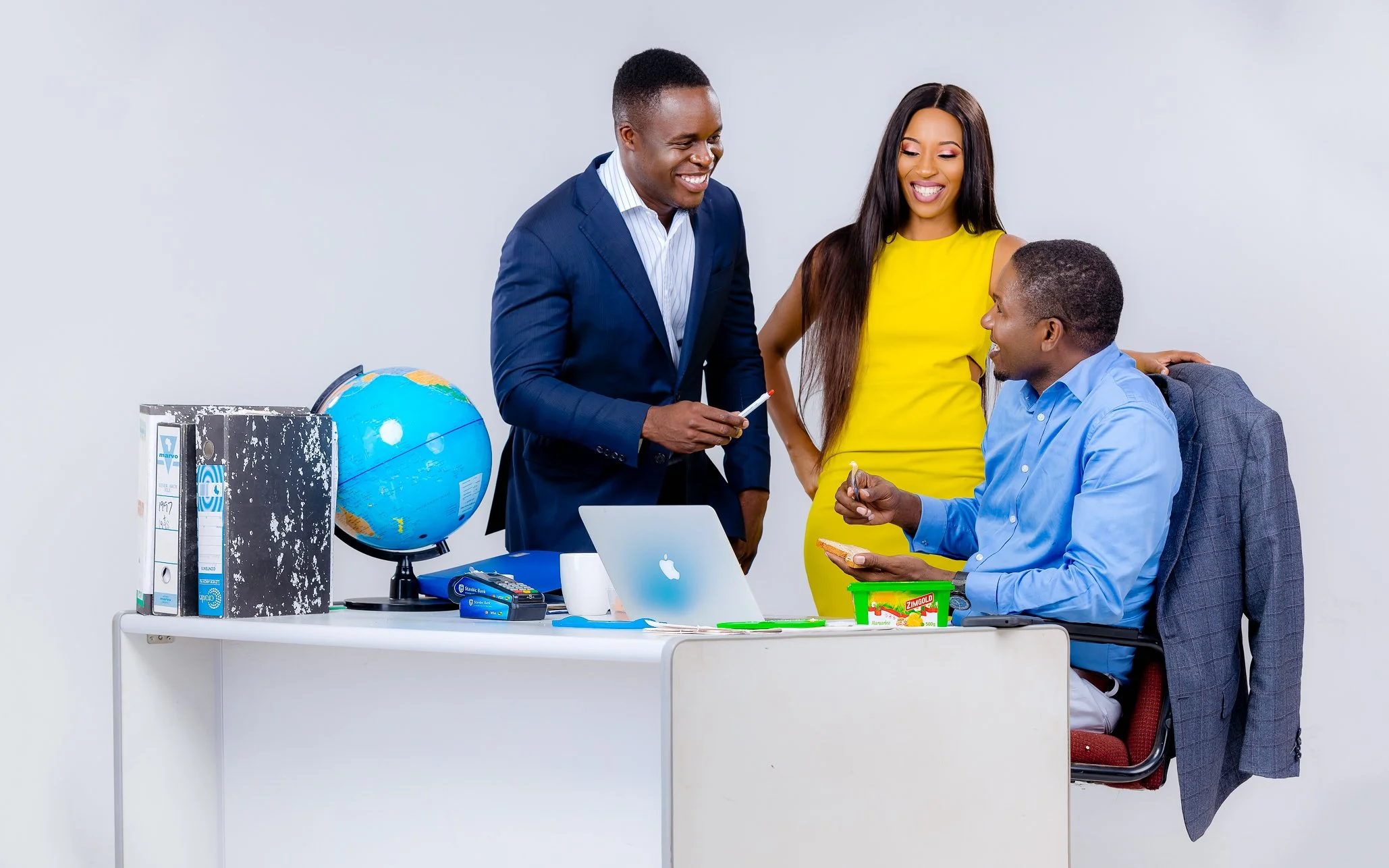 Three colleagues in an office, two men and one woman, engaged in conversation around a desk with a laptop, globe, and office supplies.