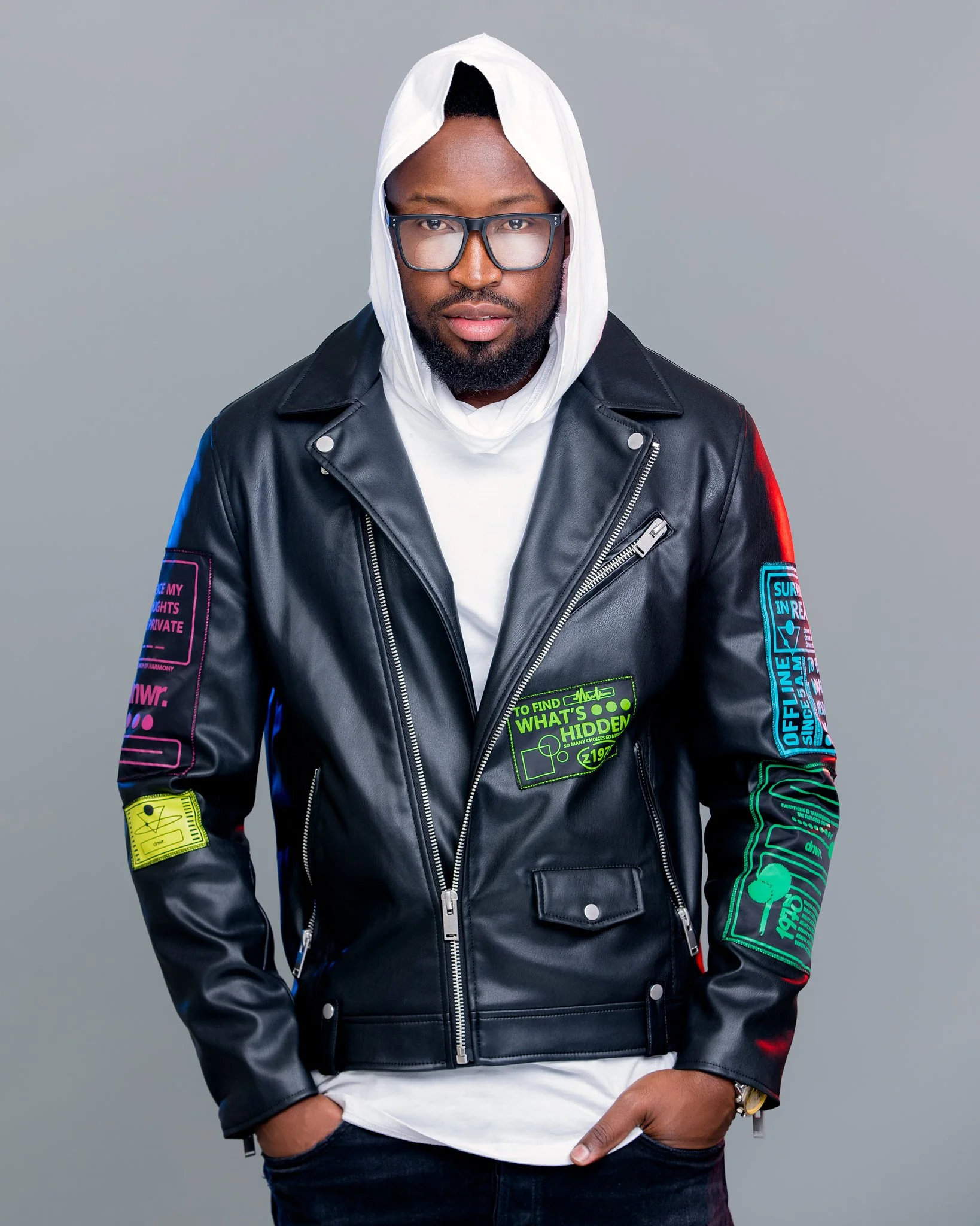 A man wearing glasses, a white hoodie, and a black leather jacket with colorful patches, standing against a gray background.