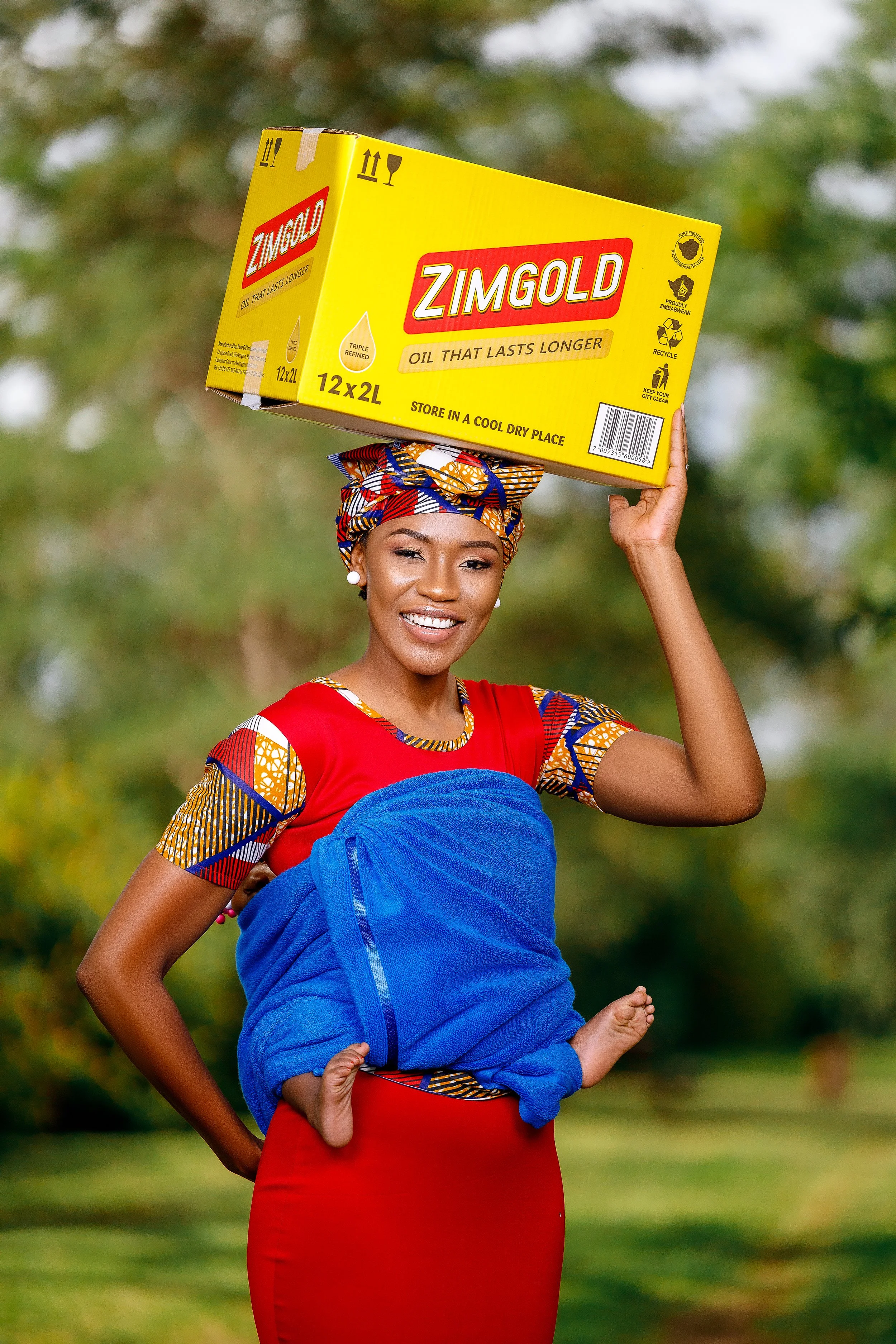 Smiling woman in a colorful outfit wearing a head wrap and holding a yellow box of Zimgold oil over her shoulder. The woman has a baby wrapped in a blue towel on her waist, holding the baby with her left hand.