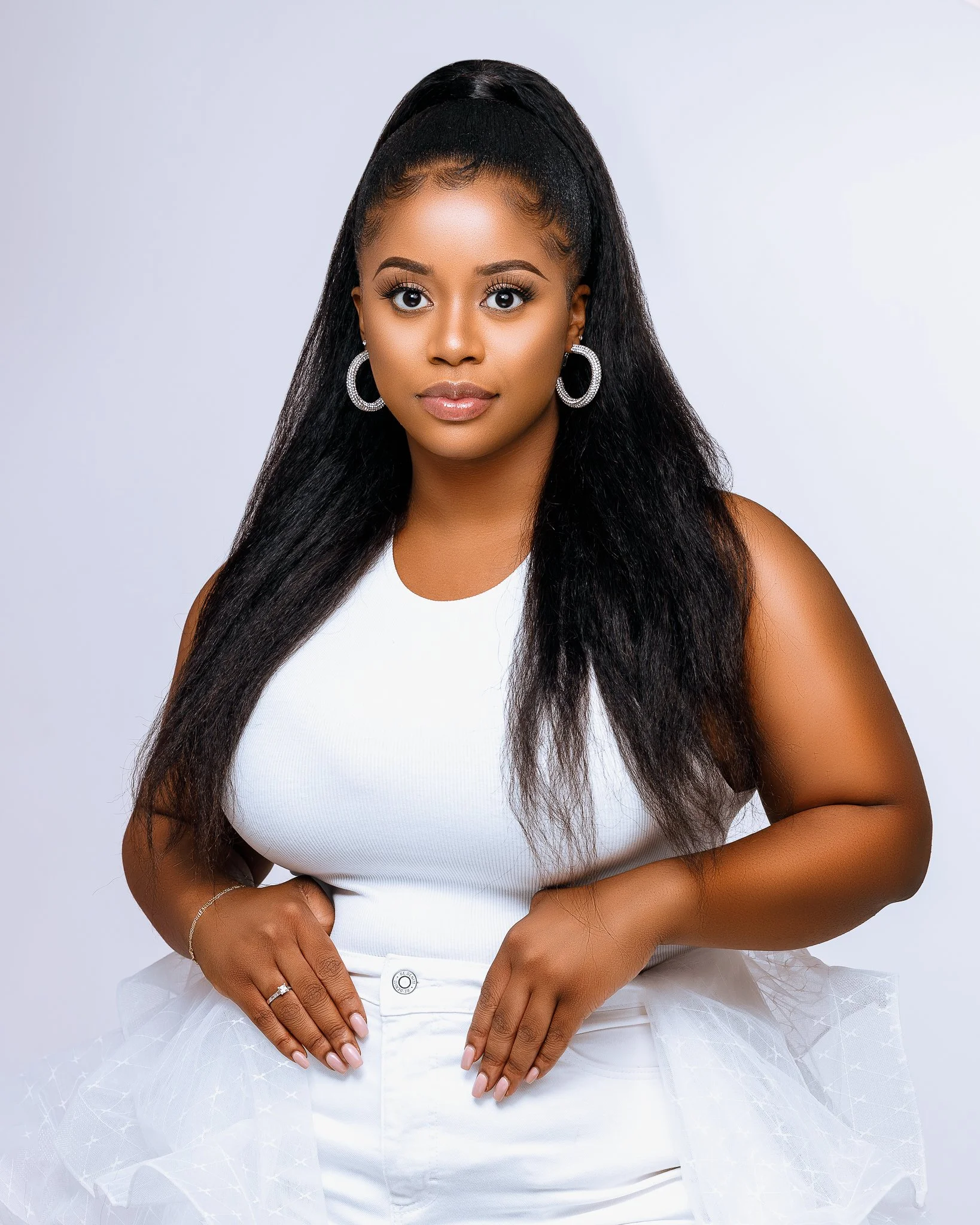 A young woman with long black hair styled in a high ponytail, wearing a white sleeveless top and white pants, standing against a light background.
