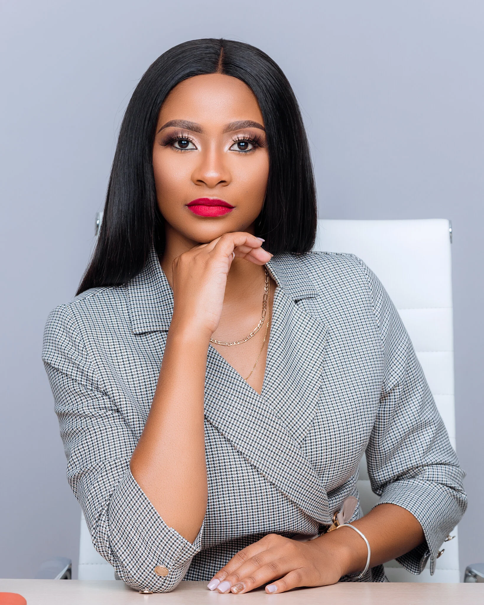 A woman with long black hair, red lipstick, and a checkered blazer sitting in a white chair against a gray background.
