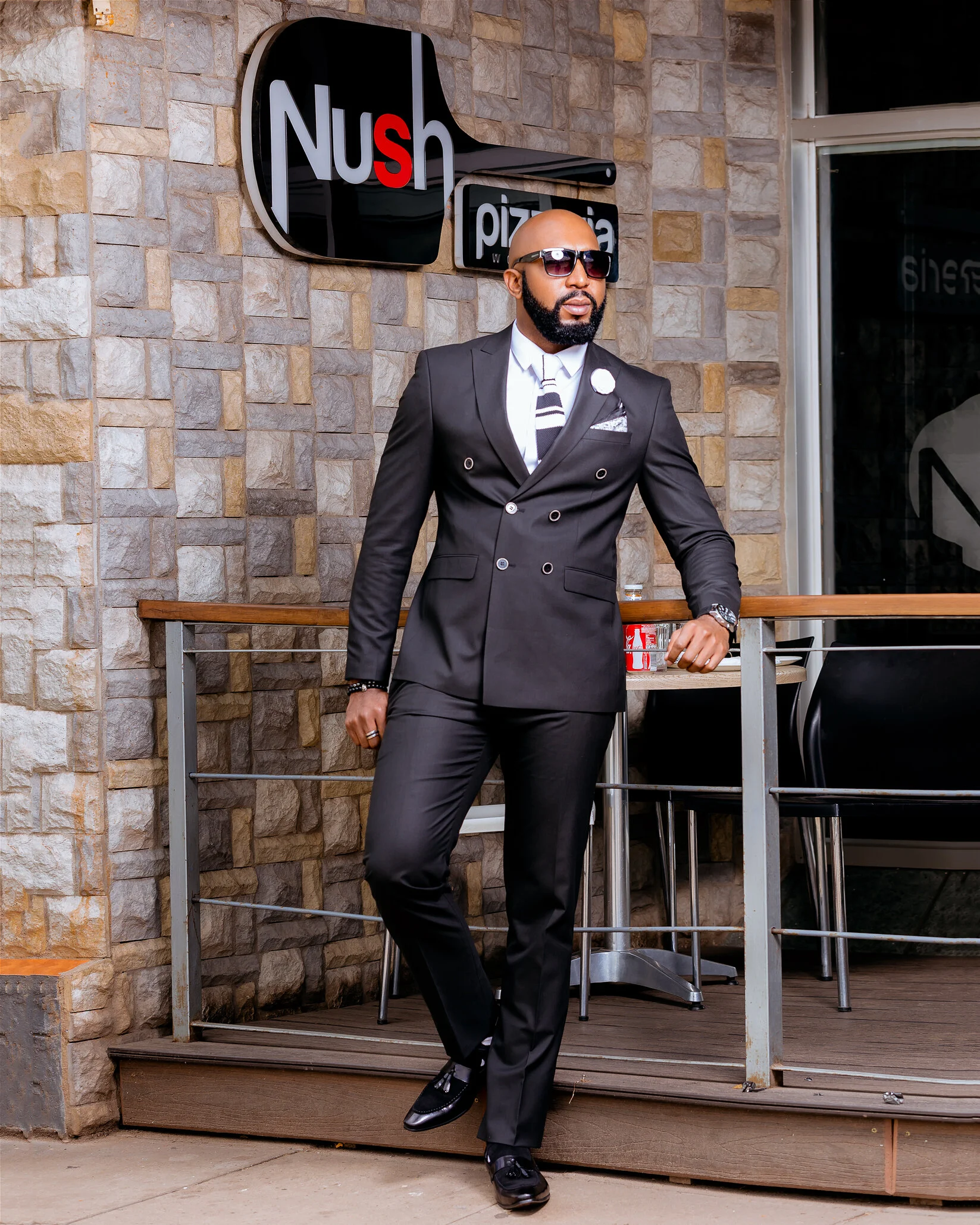 A man in a black suit, white shirt, and patterned tie posing outside a pizza restaurant, wearing sunglasses, a watch, and a pin on his lapel.