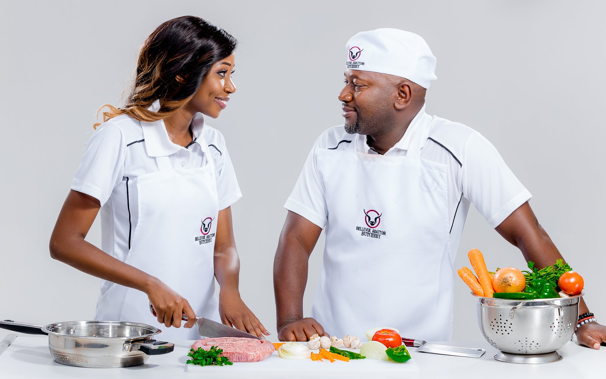 A woman and man dressed in white aprons and chef hats, smiling at each other in a kitchen, preparing vegetables and meat on a white counter.