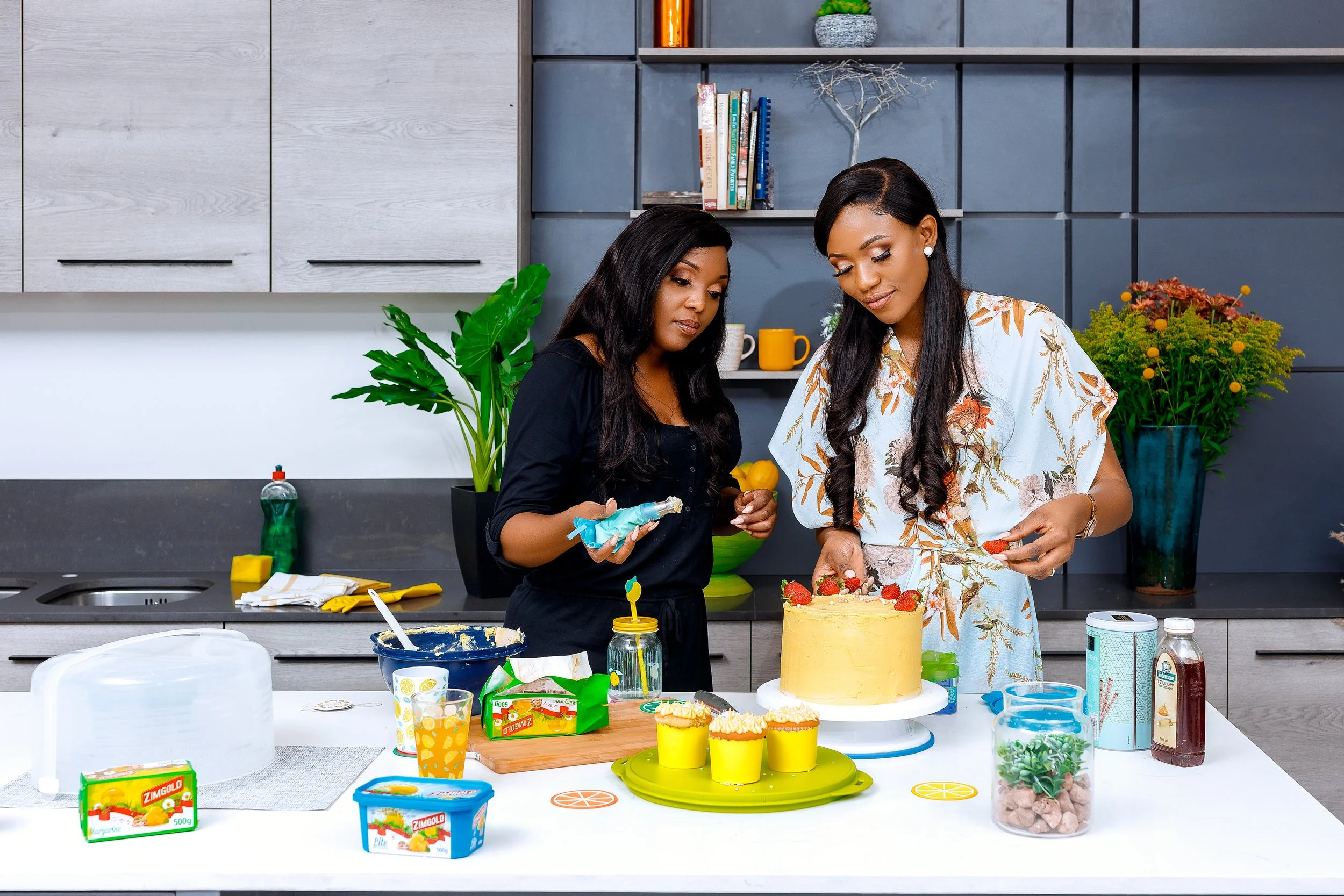 Two women in a modern kitchen decorating a birthday cake with strawberries, surrounded by cupcakes, ice cream, and baking supplies.