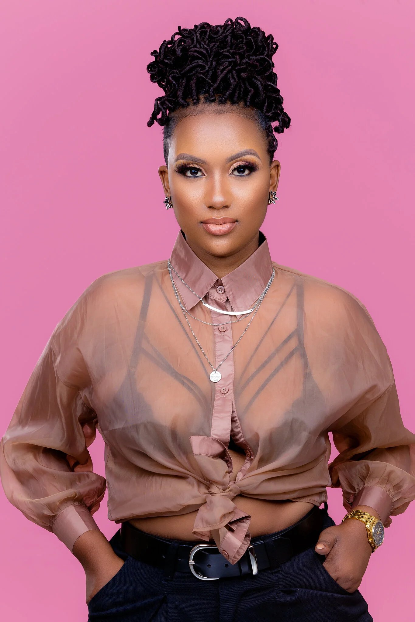 Portrait of a woman with styled black hair tied up, wearing a sheer bronze blouse tied at the waist, black pants, a black belt, and gold accessories, standing against a pink background.