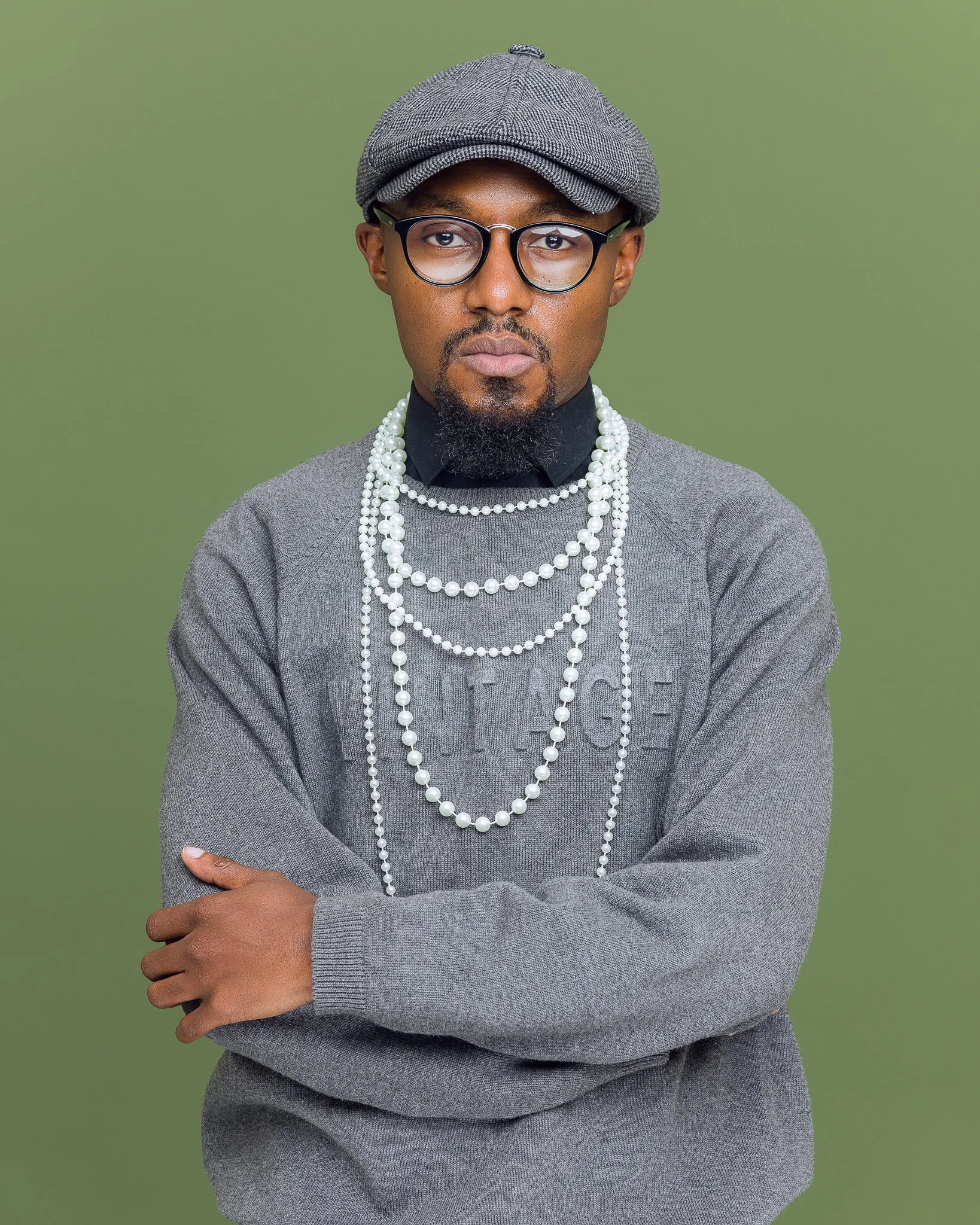A man with glasses, a grey cap, and a beard, wearing a grey sweater and multiple strands of pearls around his neck, standing against a green background.