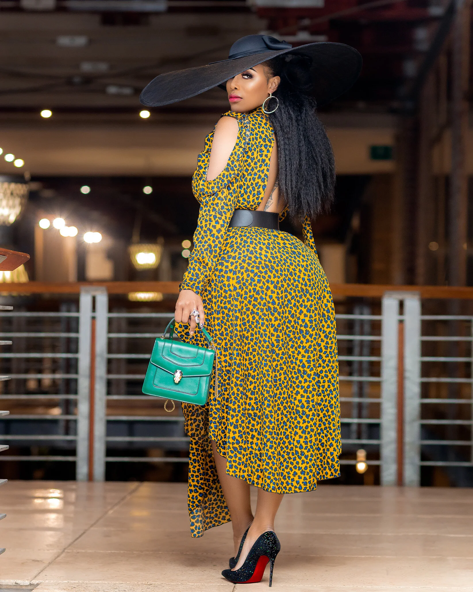 A woman wearing a large black sun hat, a yellow and blue leopard print dress with cutout shoulders, black high heels, and holding a green handbag, standing indoors with a blurred background.
