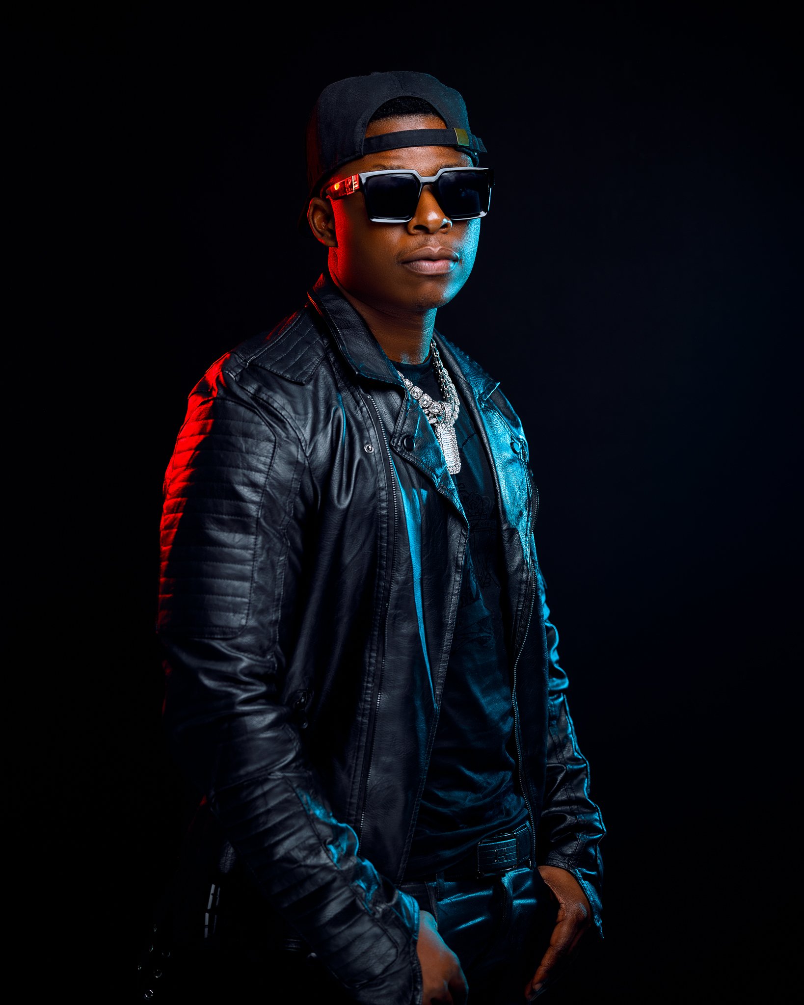 A young man wearing a black leather jacket, large sunglasses, a backwards black cap, a chunky chain necklace, and dark clothing stands against a black background with colorful lighting highlights.