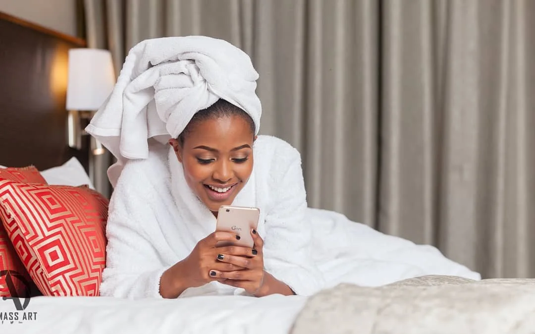 A woman with a towel wrapped around her head, lying in bed, smiling and looking at her phone.
