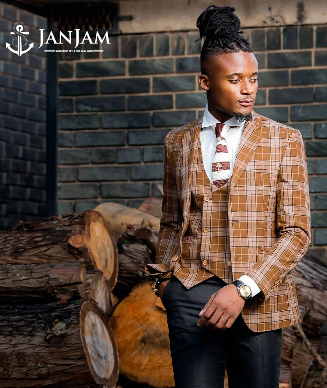 A man in a brown checkered suit with a white dress shirt and a striped tie standing outdoors near a stone fireplace with stacked firewood, against a dark brick wall.