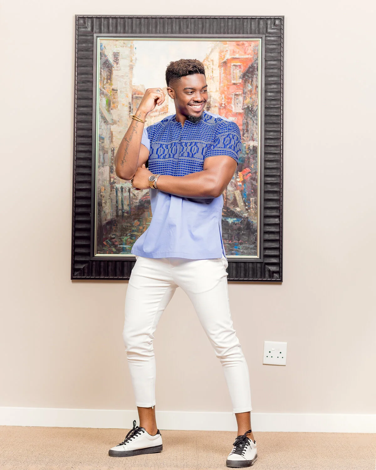 A smiling young man with a tattoo on his left arm, wearing a blue patterned shirt, white pants, and sneakers, standing in front of a colorful cityscape painting on a beige wall.