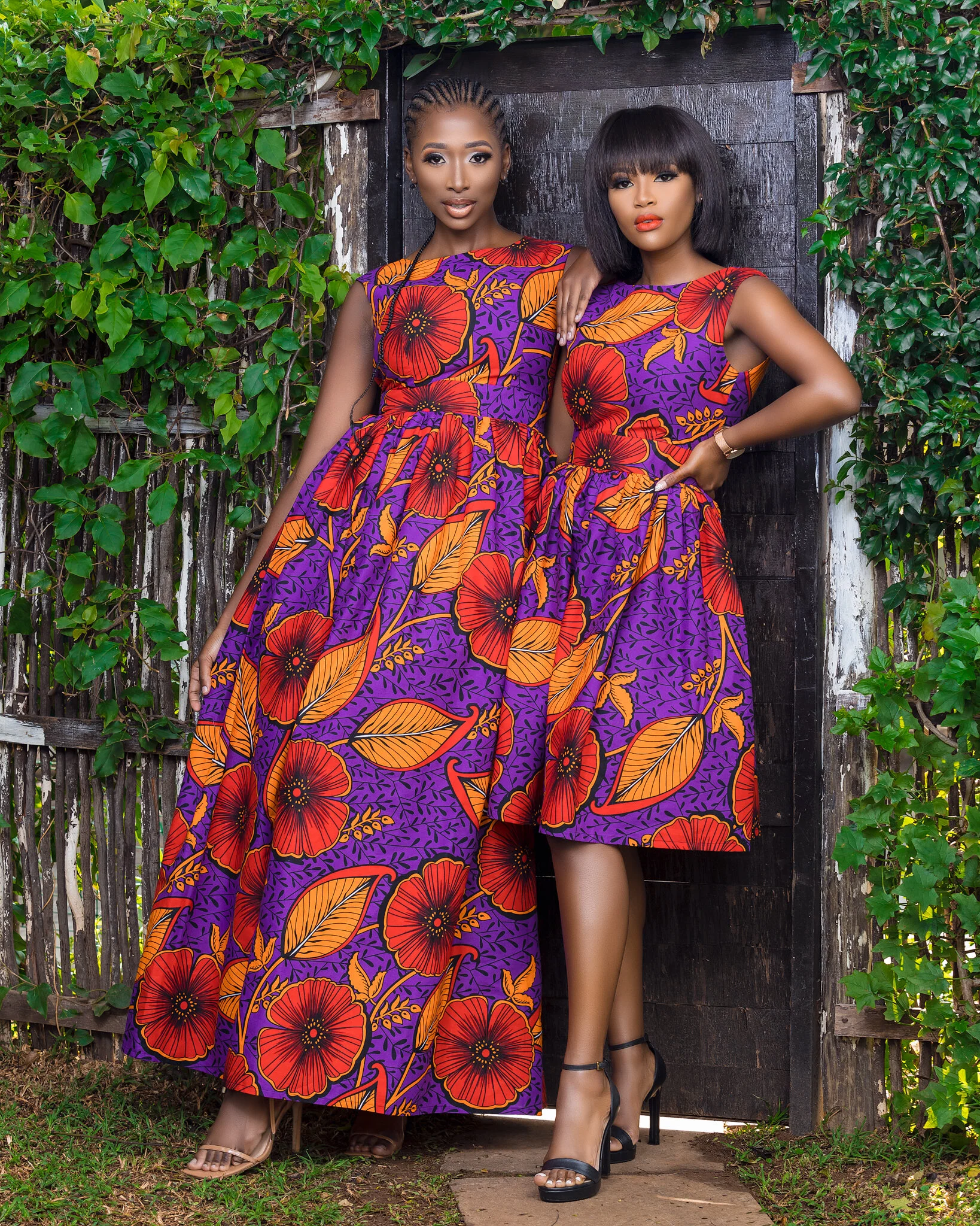 Two women wearing matching purple dresses with bright red and orange floral patterns standing outdoors in front of a black wooden door and green foliage.