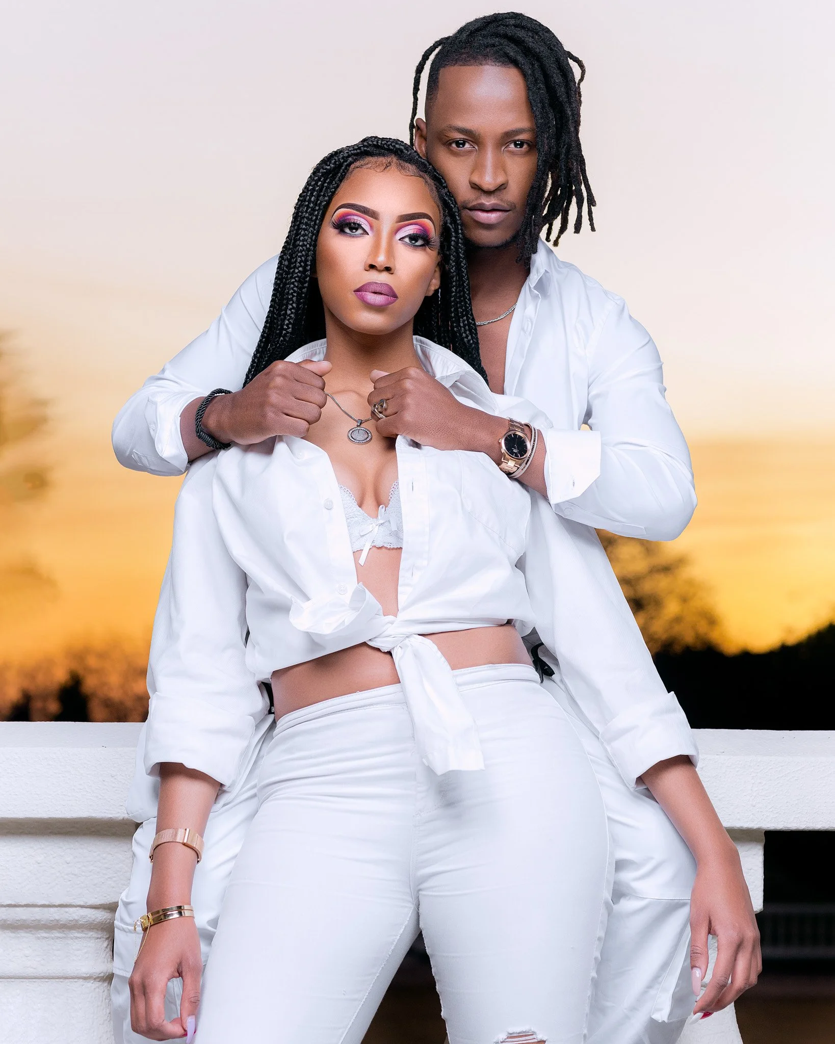A young couple dressed in white standing close together on a balcony against a sunset background. The woman has long braided hair and bold makeup, and the man has short dreadlocks. They look directly at the camera.