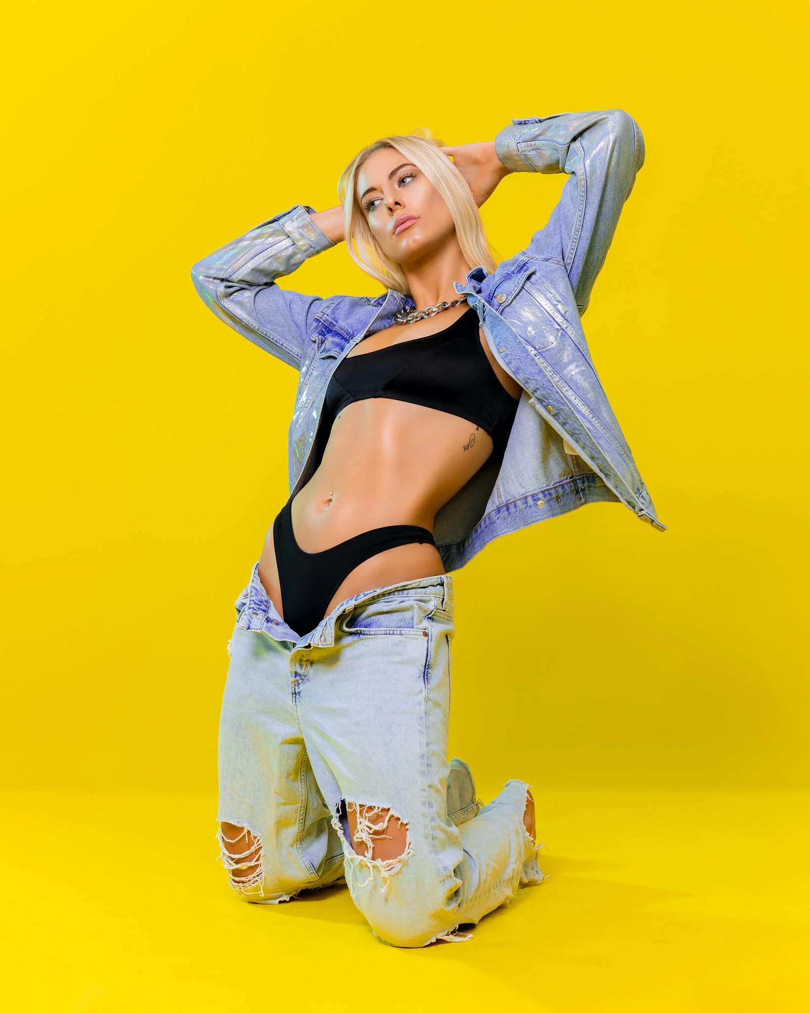 A woman with blonde hair kneeling against a bright yellow background. She is wearing a black crop top, ripped jeans, and an iridescent denim jacket, posing with her hands behind her head.