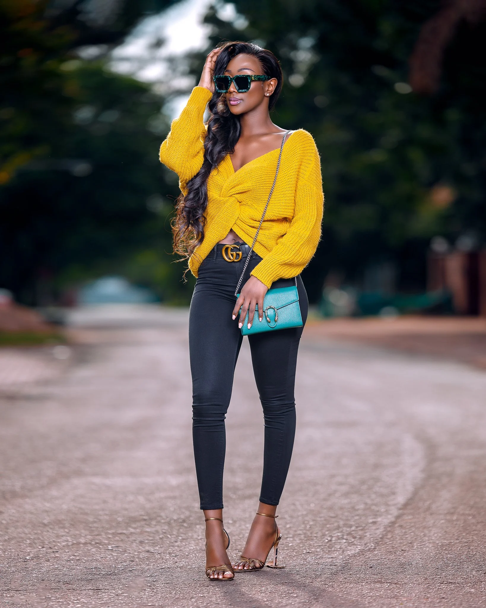 A stylish woman standing on a quiet street wearing a yellow sweater, black pants, high heels, large sunglasses, and carrying a small turquoise purse.