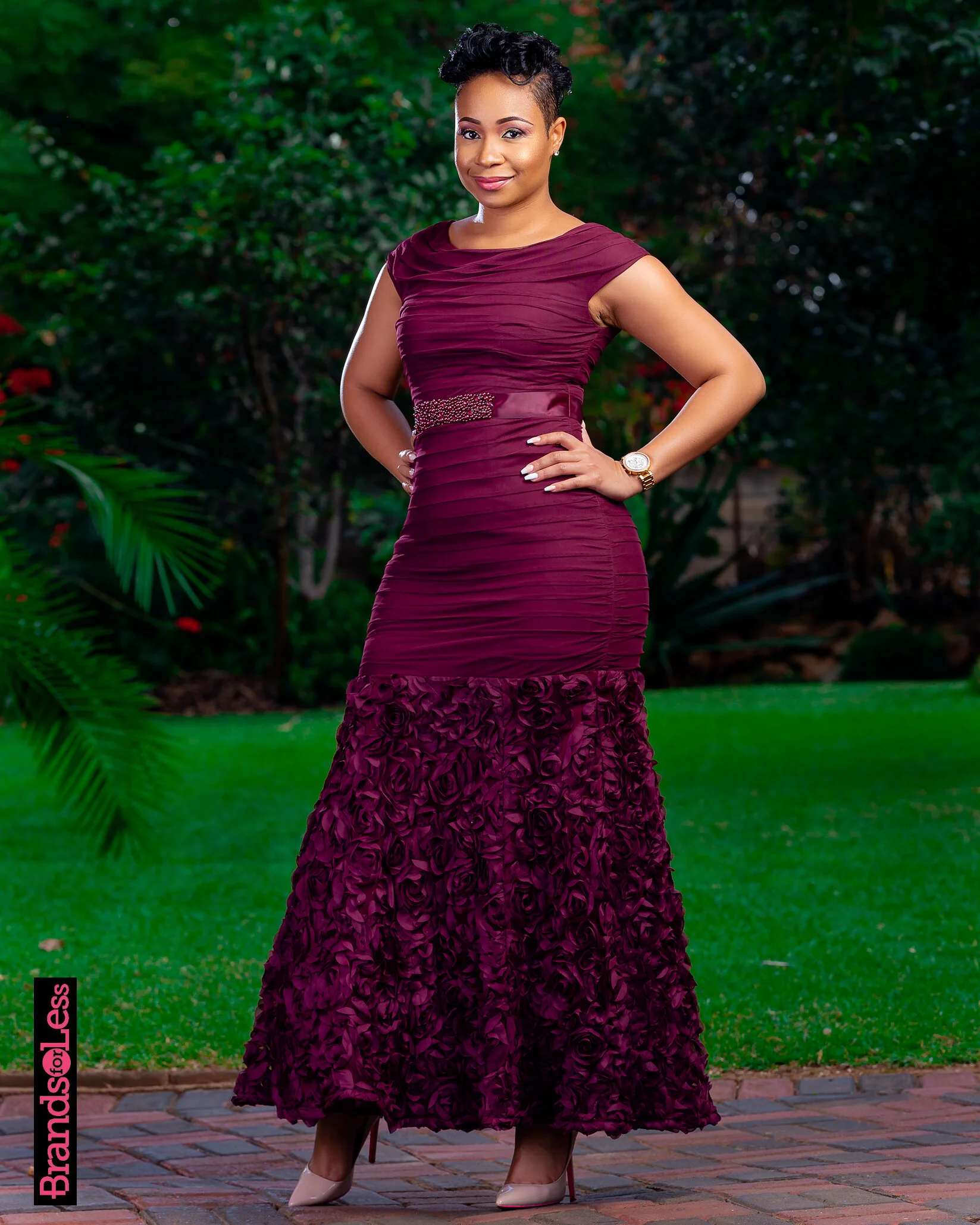 A woman in a formal burgundy evening gown with ruching details and floral embellishments at the hem, standing outdoors on a brick pathway with greenery in the background.
