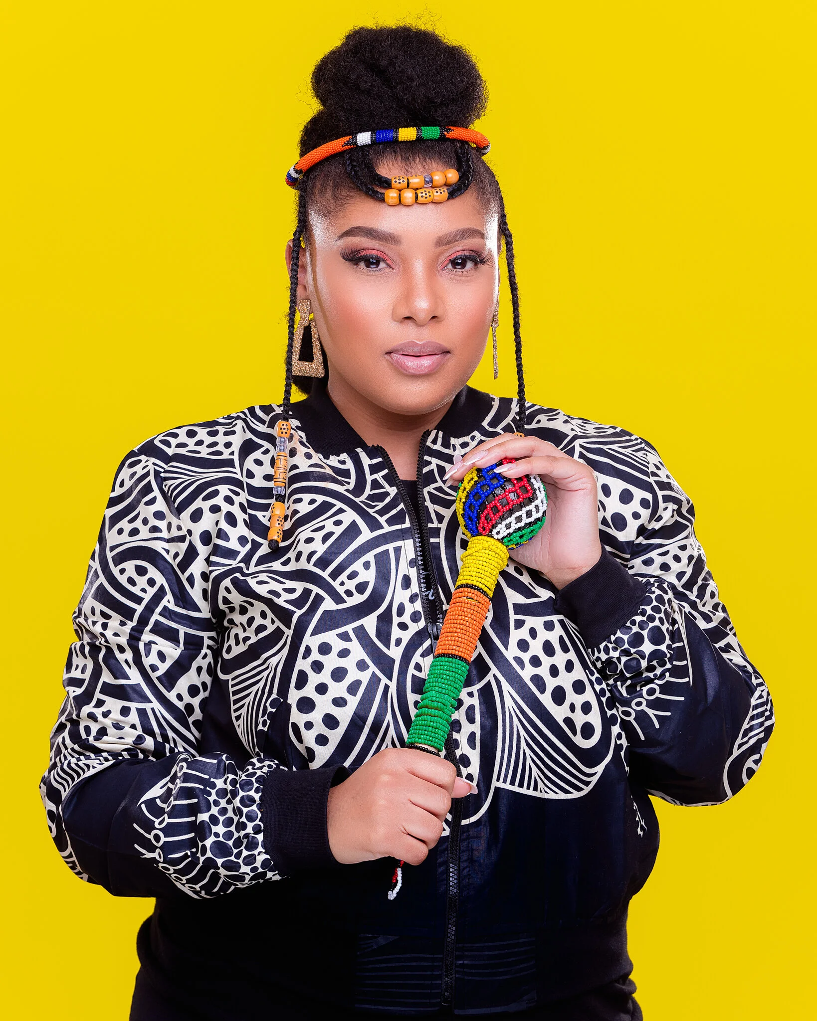 A woman with dark skin, braided hair with beaded jewelry, wearing a black and white patterned jacket, holding a colorful beaded staff, standing against a bright yellow background.