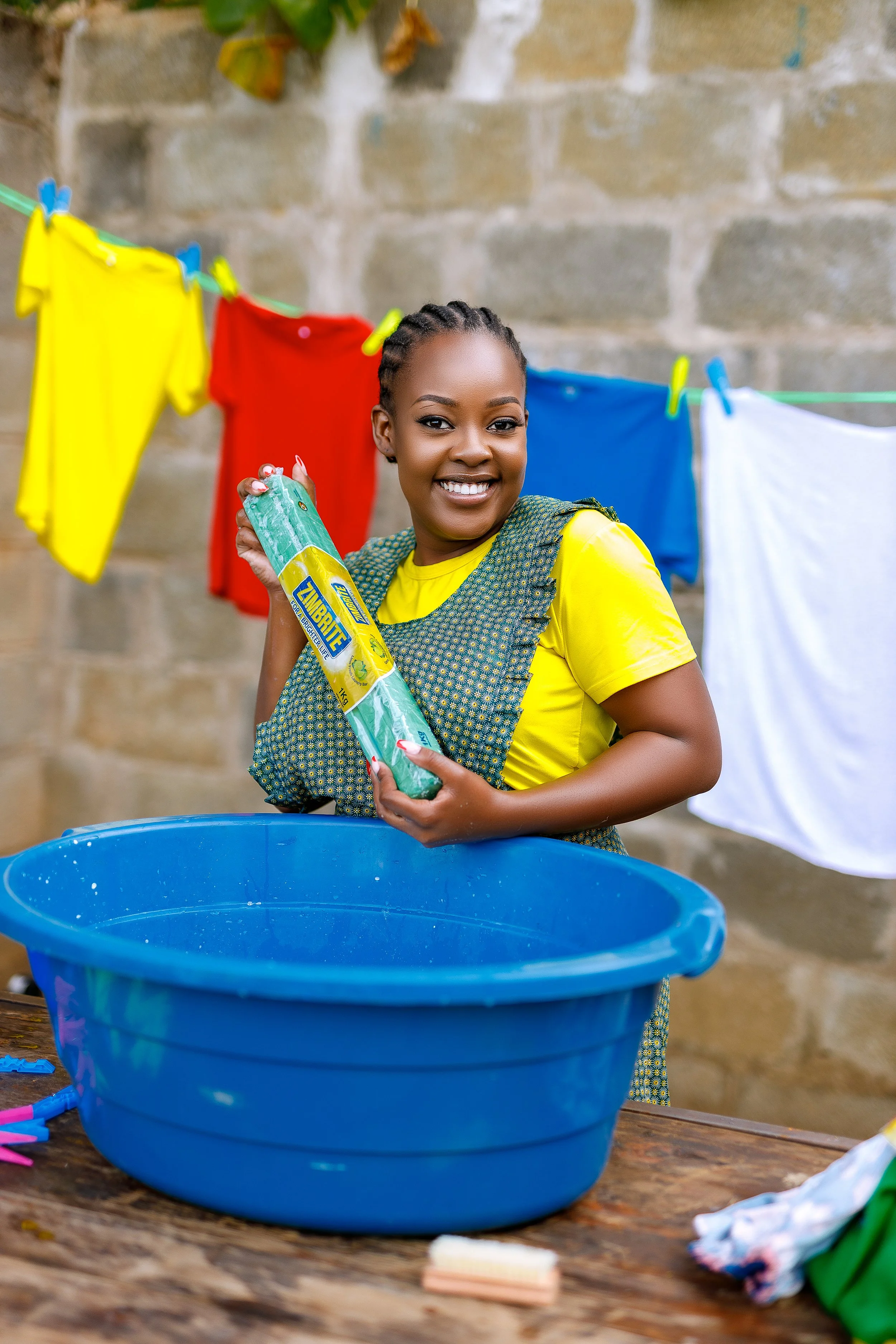 A woman smiling while doing laundry outdoors. She is holding a pack of Zorbrite bleach next to a blue wash basin. Clothes are hanging on a line behind her against a stone wall.
