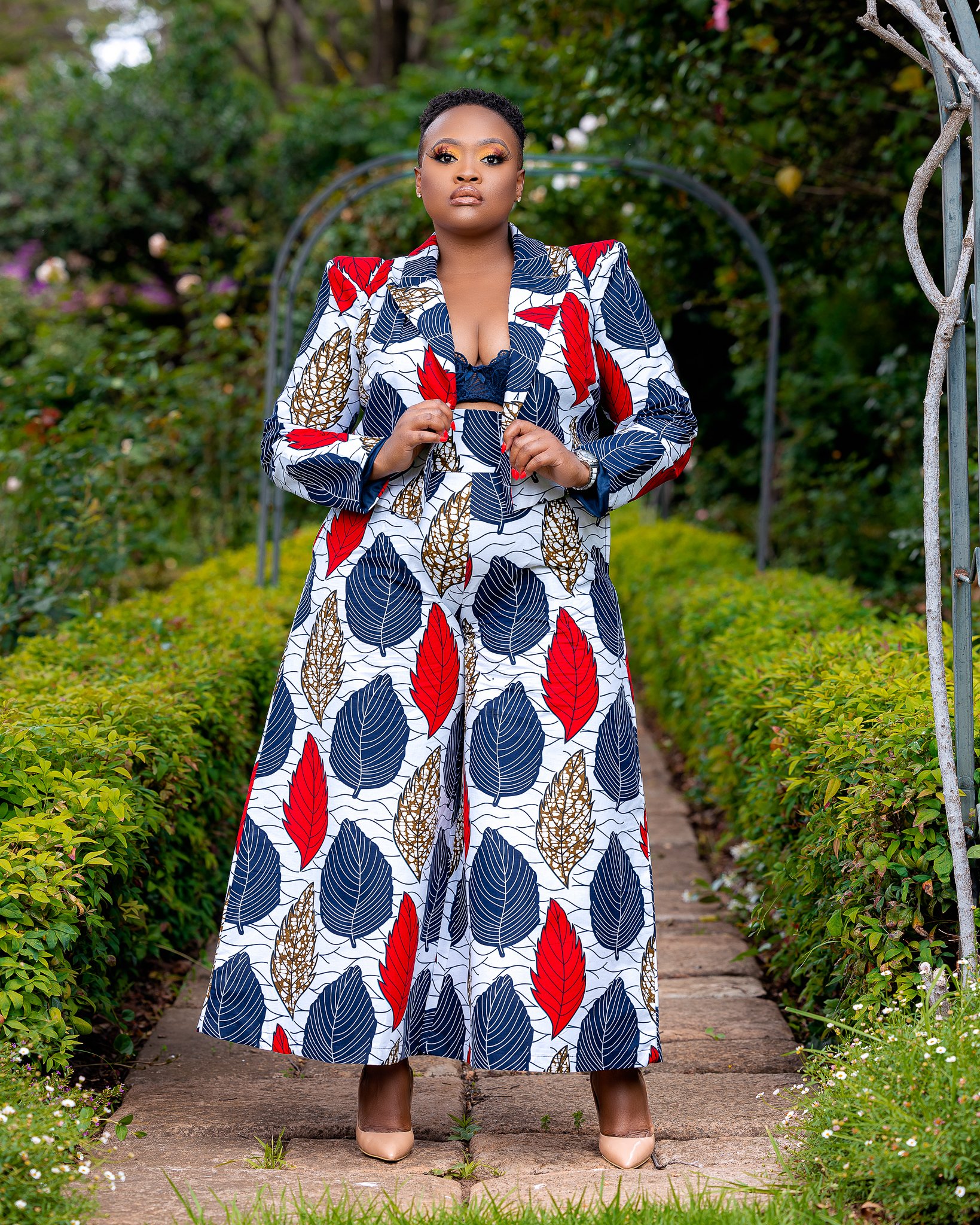 A woman stands on a stone path in a garden, wearing a matching leaf-patterned jacket and wide-leg pants with red, navy, gold, and white leaves. She has short black hair, makeup, and nude heels, with her hands holding the lapels of her jacket.