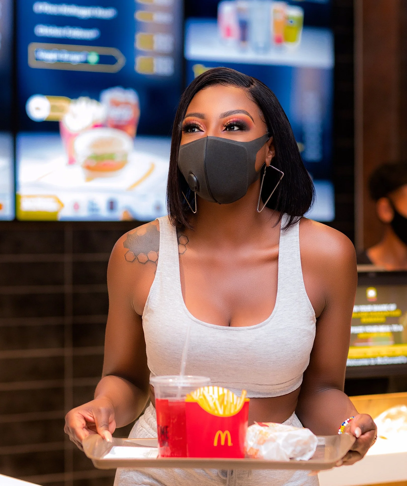 A woman in a white tank top and black face mask holding a McDonald's tray with fries and a soft drink, standing in a fast food restaurant.