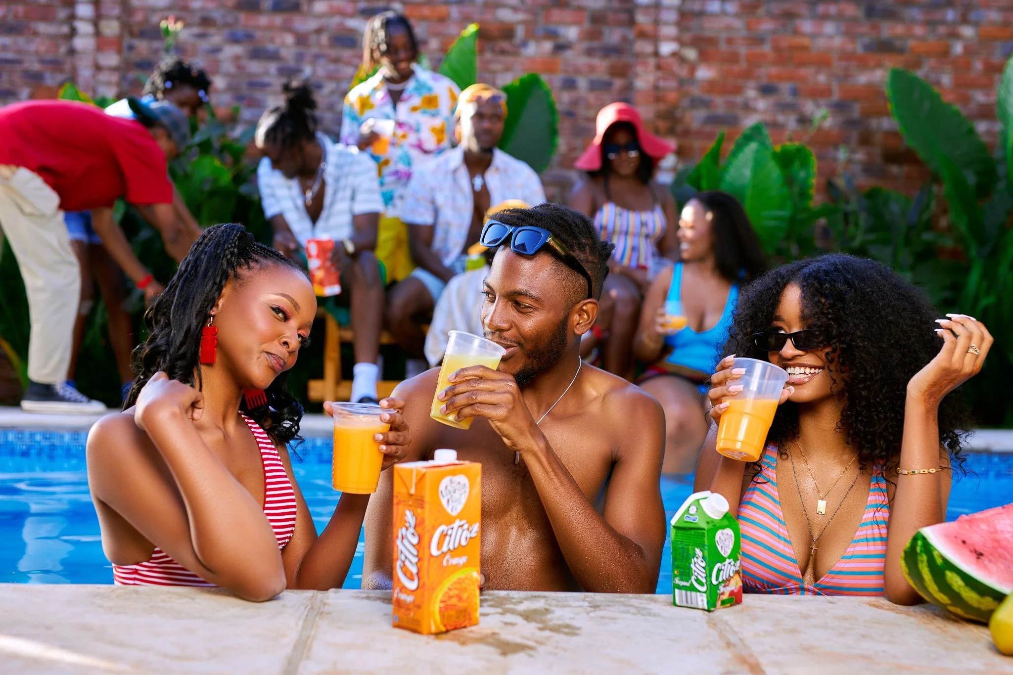 Three friends enjoying drinks by the poolside during a summer party, with a group of people relaxing on chairs and a brick wall with green plants in the background.