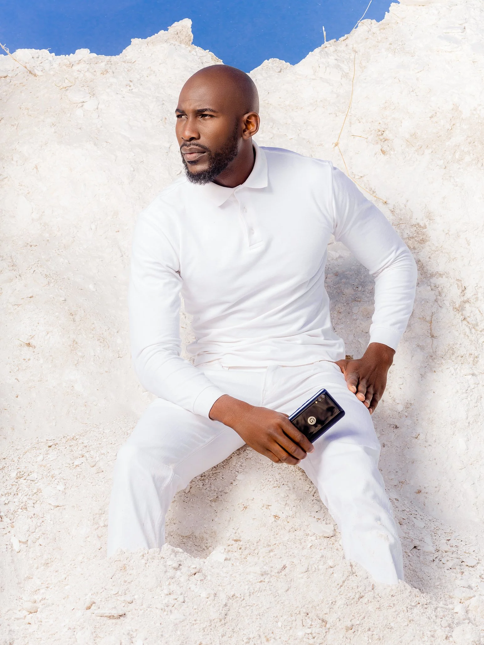 Man sitting on white sand holding a smartphone with a black case, dressed in white, with a dirt wall and blue sky behind him.
