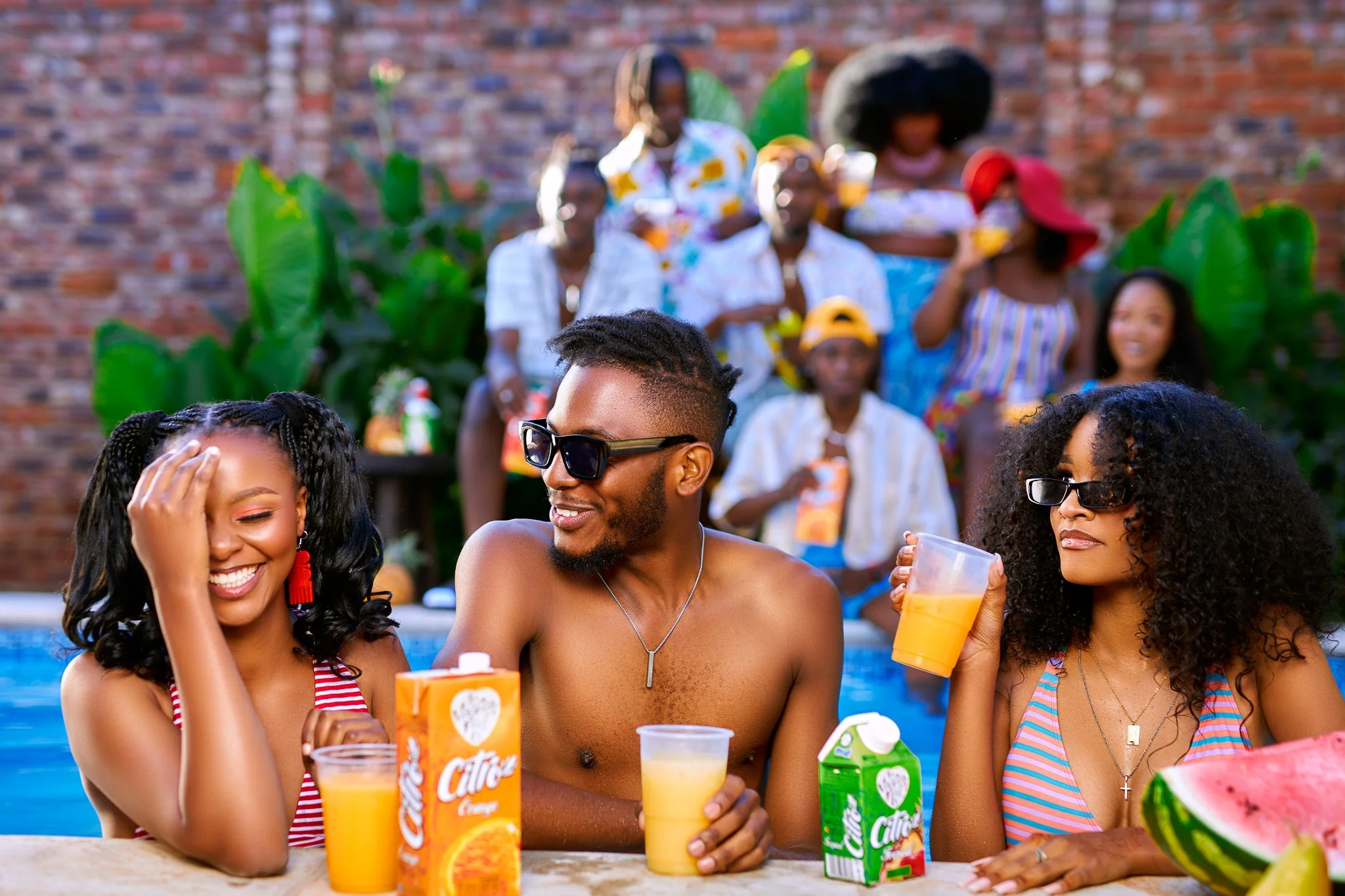 Group of friends enjoying drinks by a pool, with some wearing sunglasses and tropical drinks, in a backyard with brick wall and large green plants.
