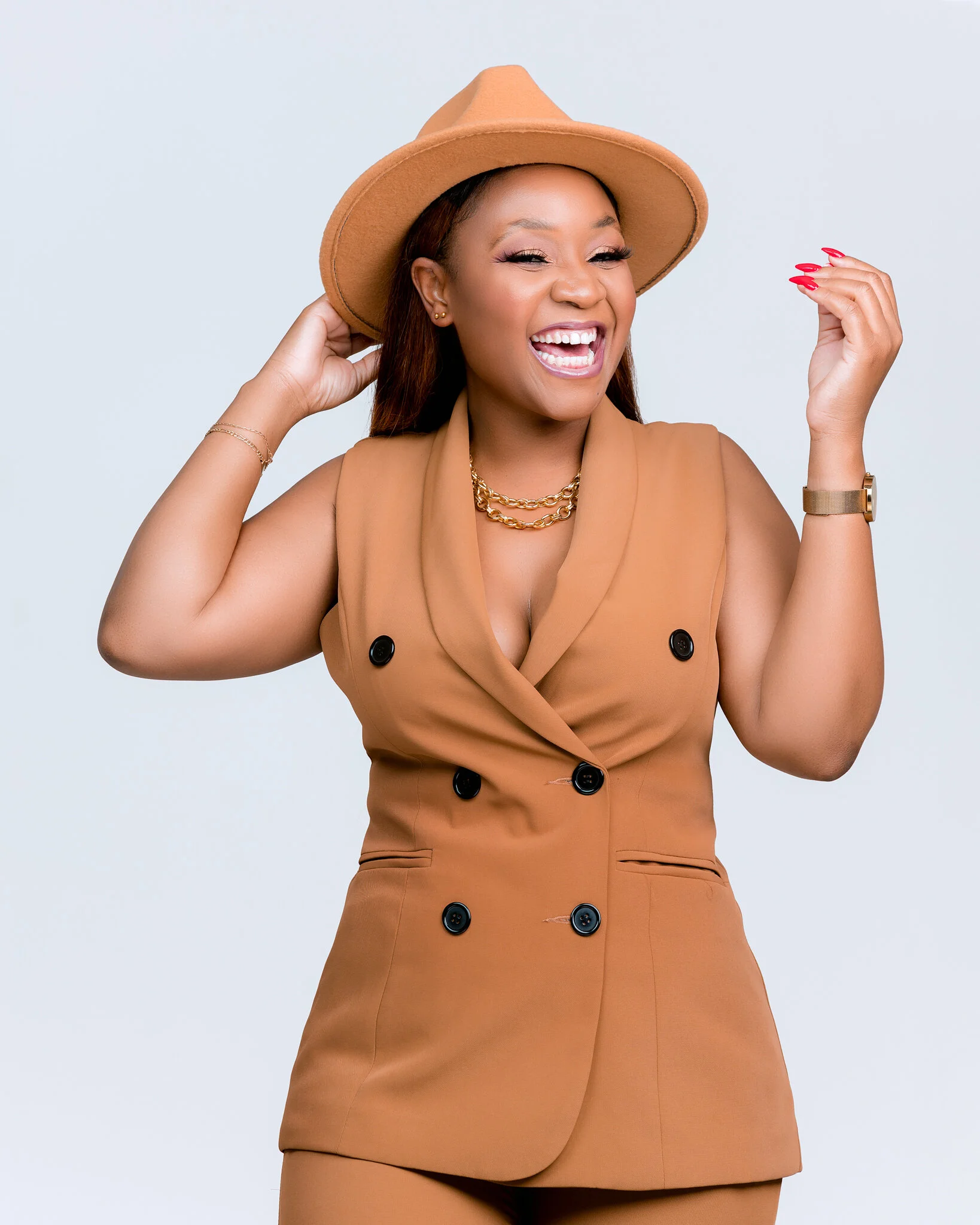 A woman in a tan sleeveless blazer with black buttons, wearing a matching tan wide-brimmed hat, smiling and posing with one hand on her head and the other slightly raised. She is accessorized with gold jewelry including layered necklaces, earrings, a