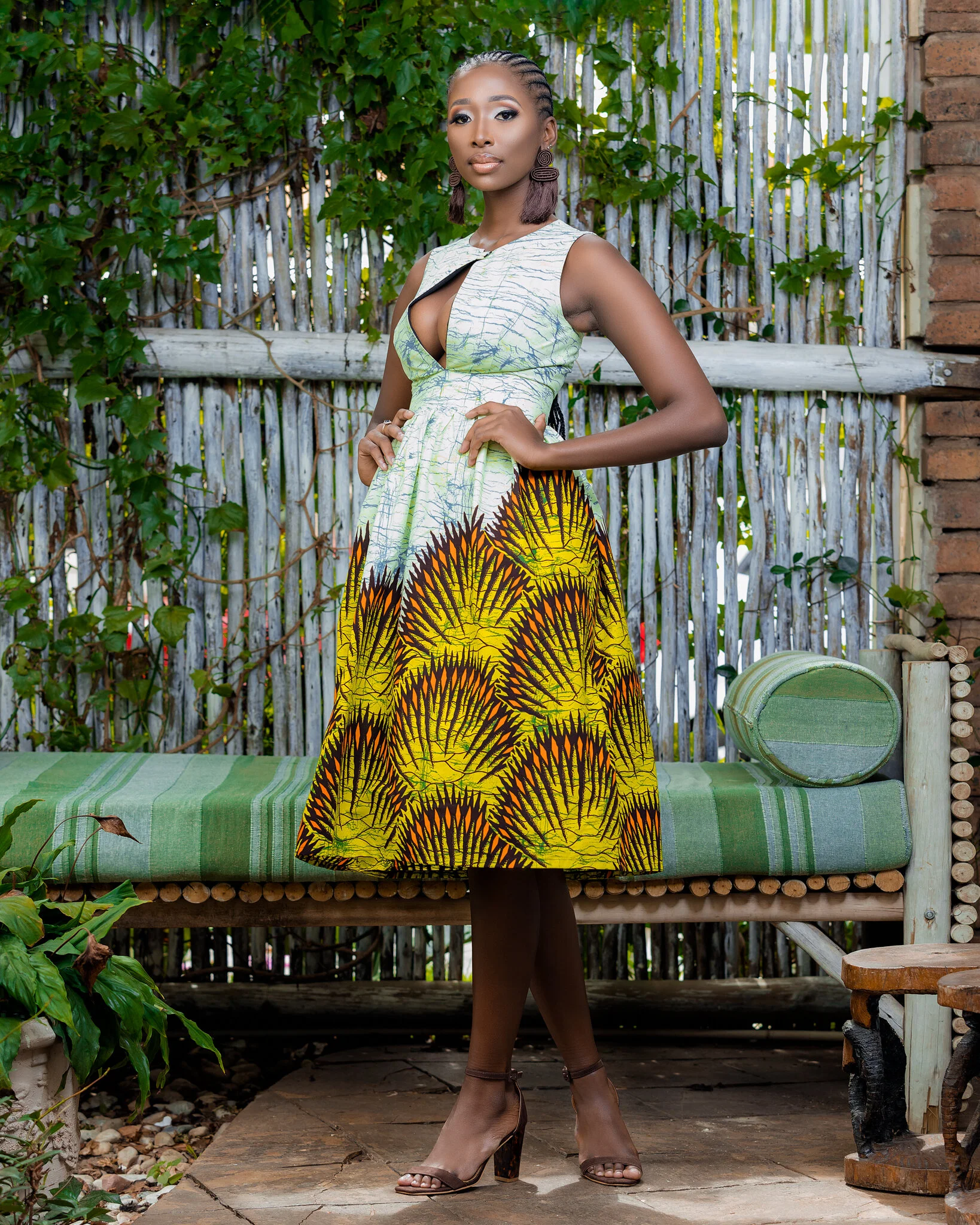 A woman in a colorful dress standing outdoors on a wooden floor, with a bamboo and leaf background, near a wooden bench with green striped cushions.