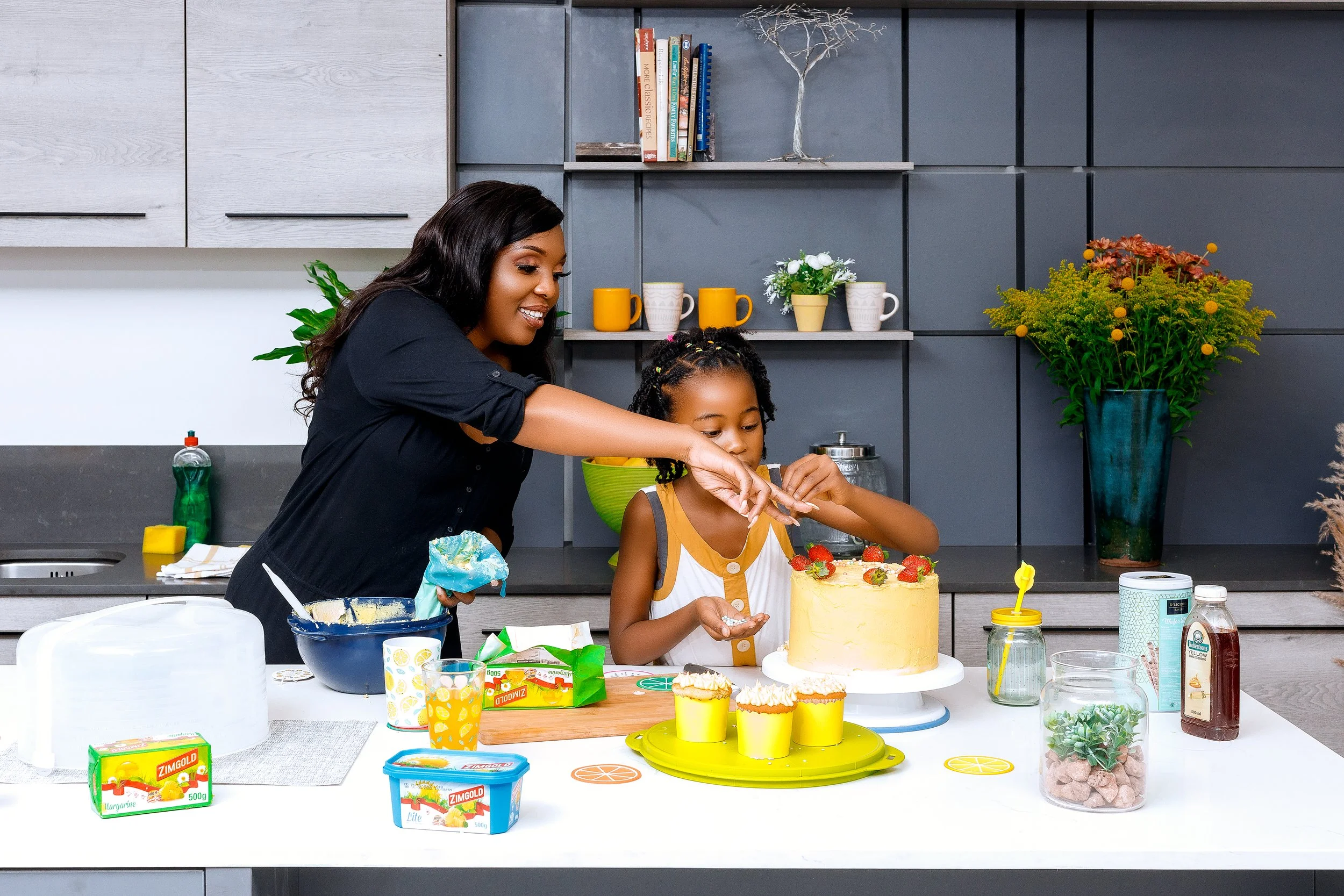 A woman and a young girl decorating a yellow birthday cake in a modern kitchen surrounded by various baking ingredients and utensils.
