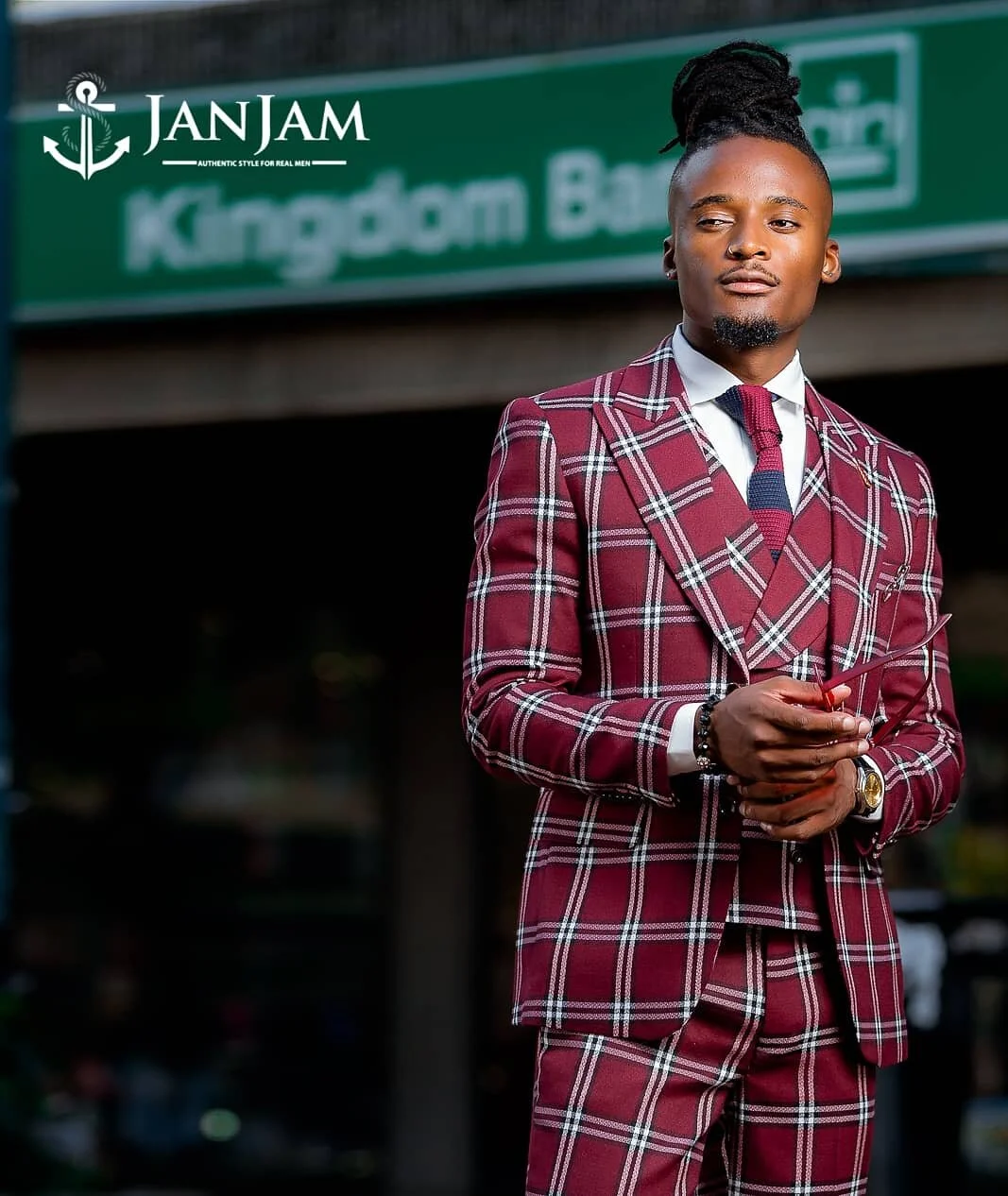 A young man standing outdoors in front of a green sign with the text 'Kingdom Bank'. He has long dreadlocks styled in a bun, is dressed in a red and white plaid suit with a white shirt and a dark tie, and is holding a red mobile phone.