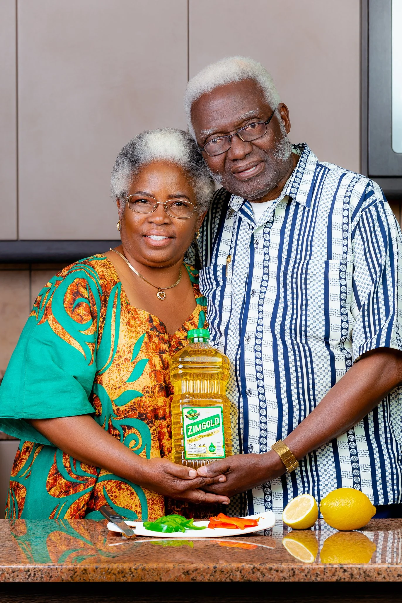An elderly African American couple standing in a kitchen, holding a bottle of Zimgold soybean oil together, with a lemon, a plate of cut vegetables, and a whole lemon on the counter in front of them.