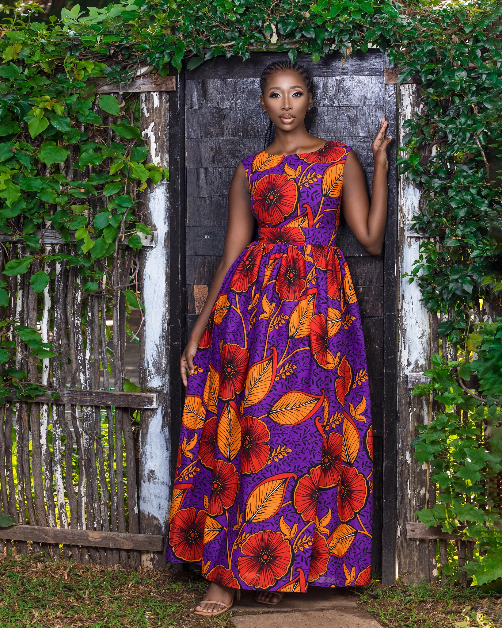 A woman in a colorful purple dress with red and orange floral patterns standing in front of a rustic wooden door surrounded by green ivy.