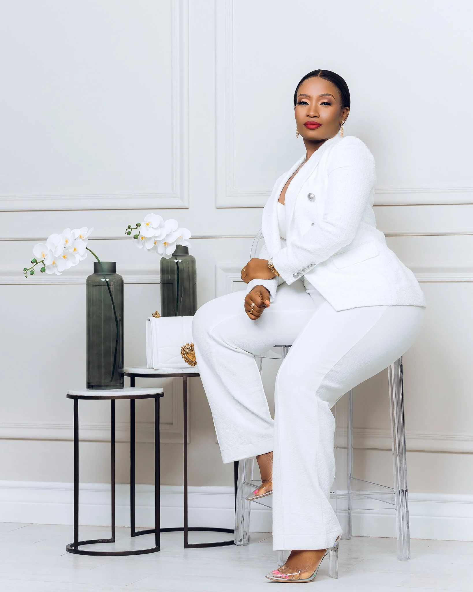 A woman in a white suit and pants sitting on a clear chair in a well-lit room with white walls, next to a small black and white table with two tall dark green vases with white orchids, and a white purse with gold embellishments.
