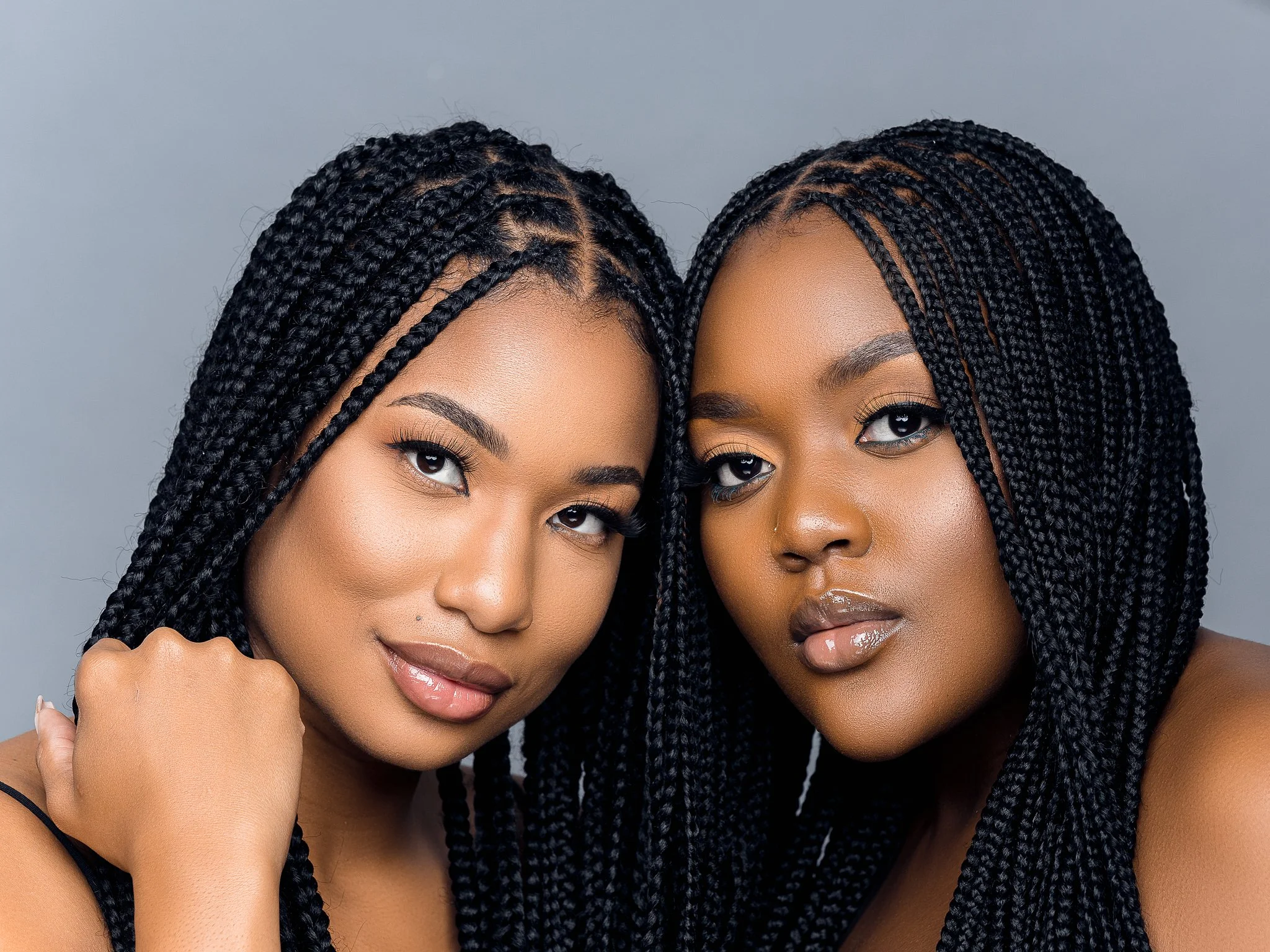 Two women with dark braided hairstyles posing closely against a gray background.
