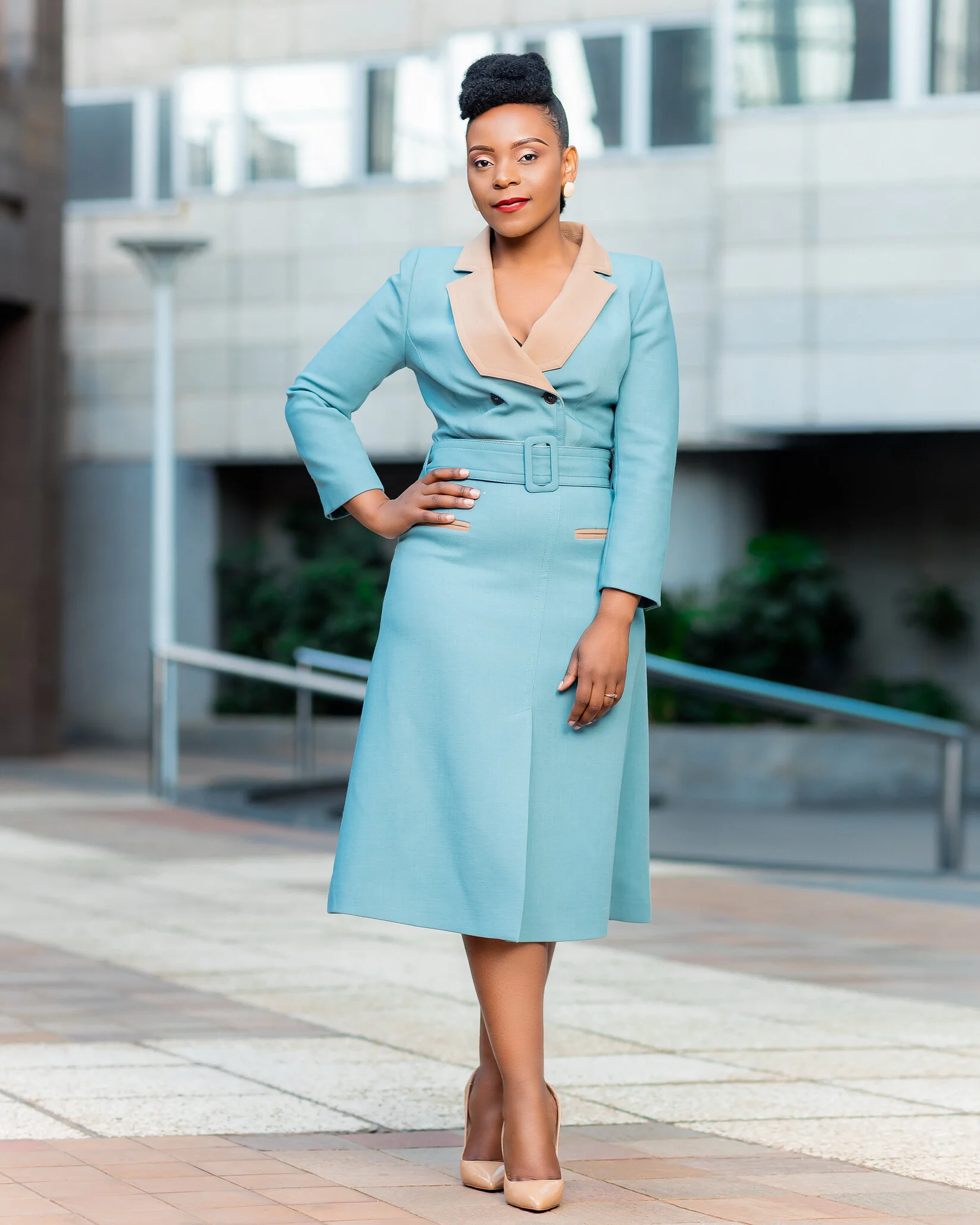 A woman in a light blue tailored trench coat dress with a beige collar and belt, standing outdoors in front of a modern building with glass windows.