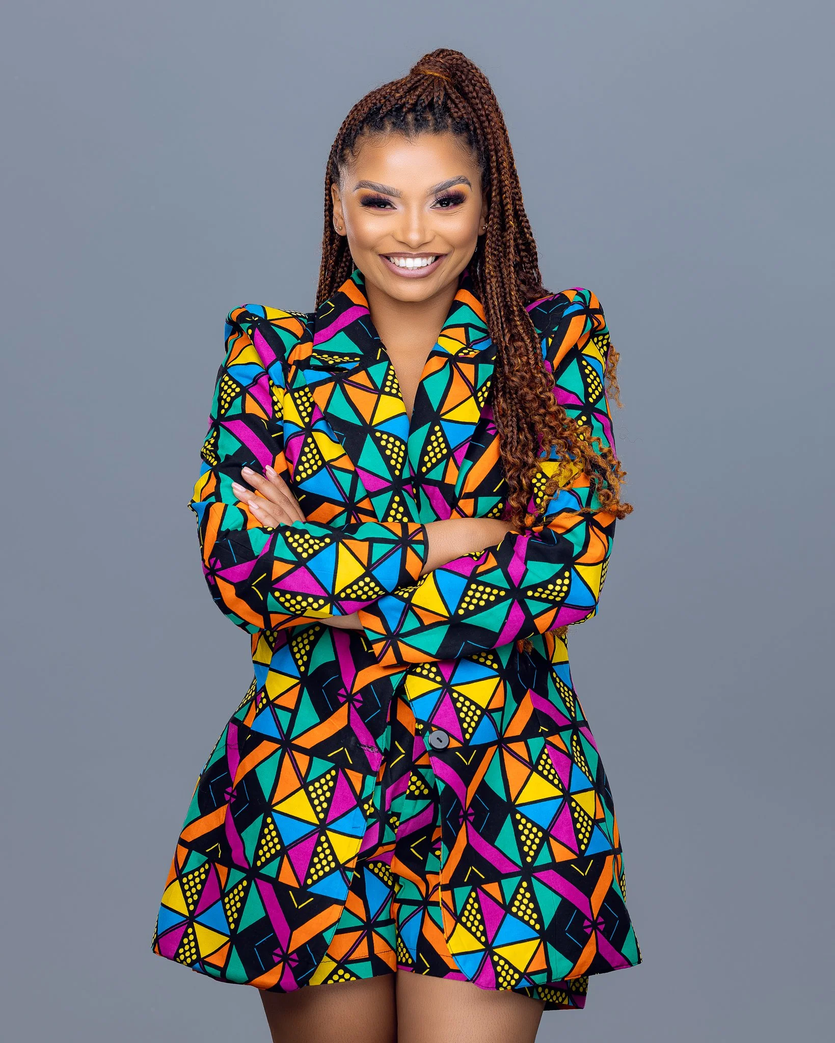 A young woman with braided hair, smiling, wearing a colorful geometric patterned blazer and matching shorts, standing against a plain gray background.