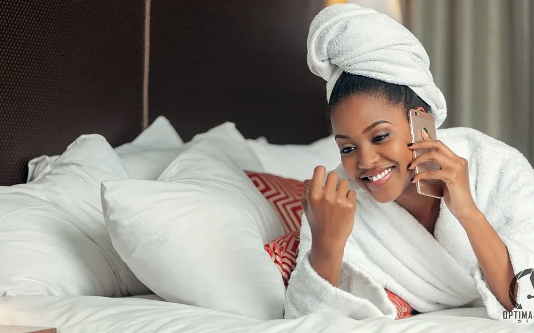 Woman smiling and talking on a phone while relaxing on bed with white pillows and a bed towel wrapped around her head.