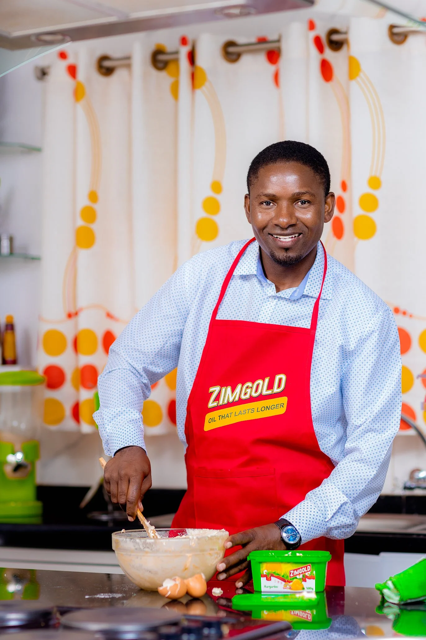 Man in a red apron stirring batter in a glass bowl in a kitchen.