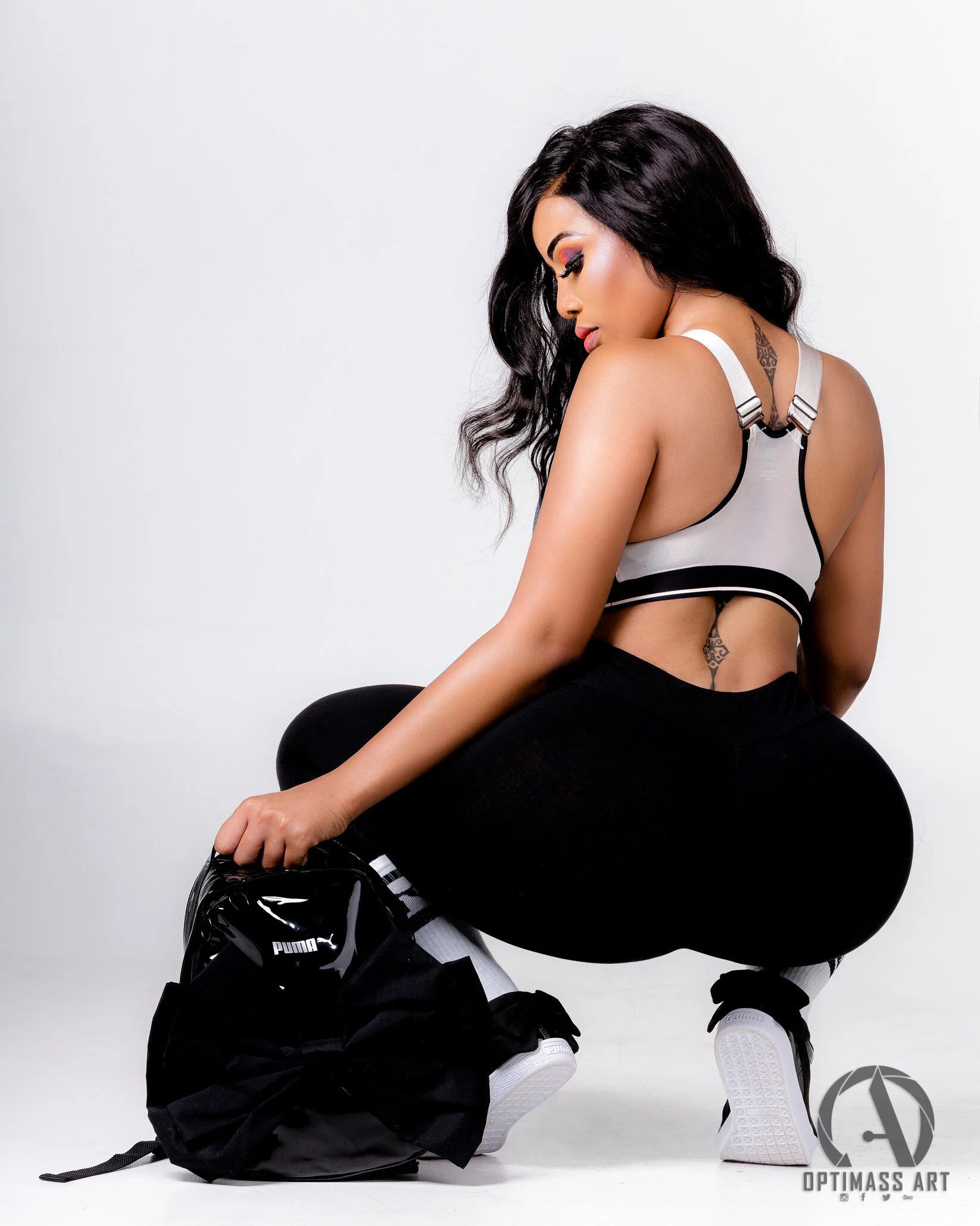 A woman with long dark hair, wearing a grey sports bra, black leggings, and white sneakers, squatting on the floor holding a black Puma backpack against a white background.