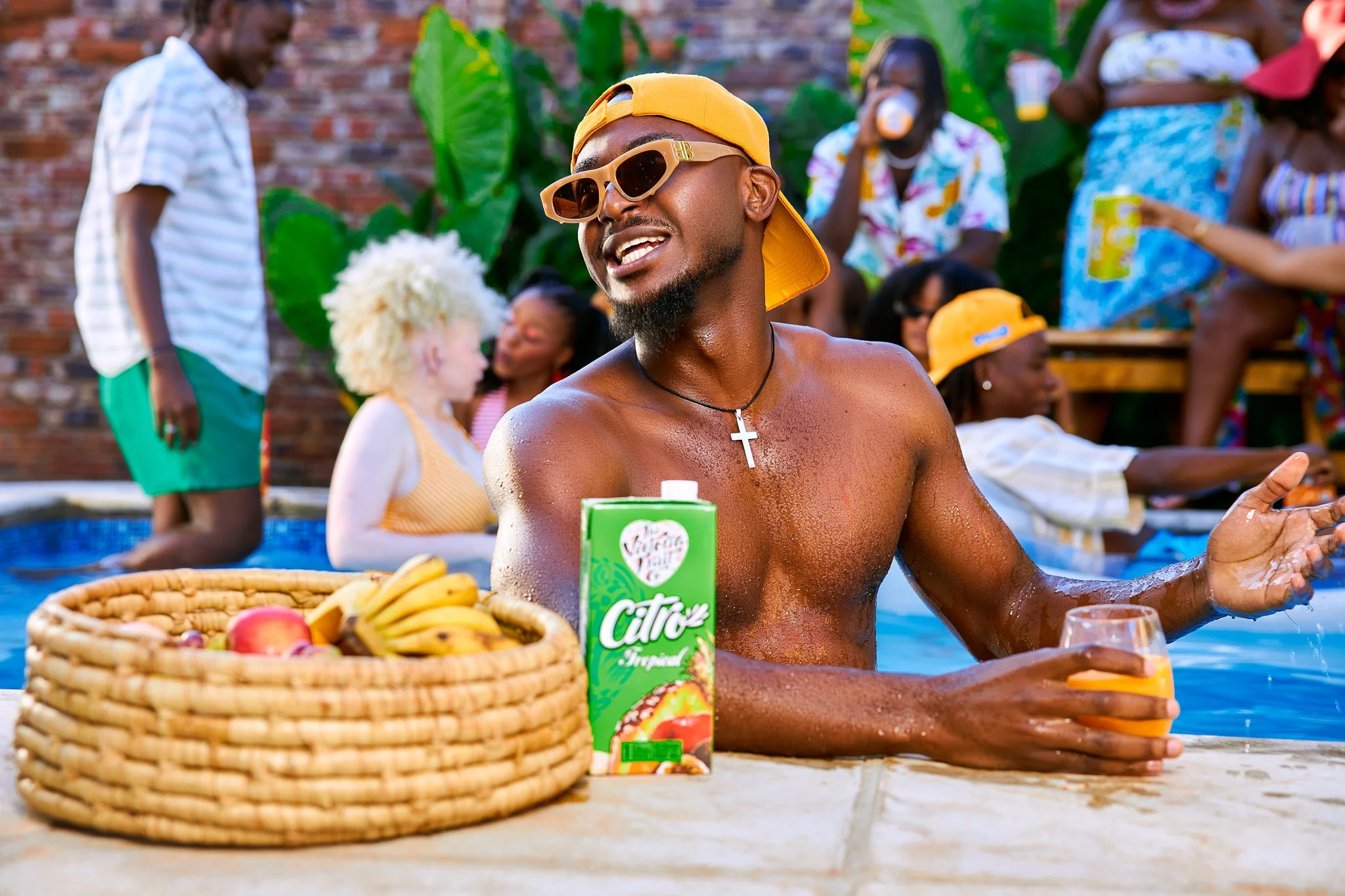 A group of people enjoying a pool party outdoors with a man in the foreground, wearing sunglasses, a yellow cap backwards, and a cross necklace, smiling and holding a glass of orange drink. There is a basket of fruit and a carton of juice in front of