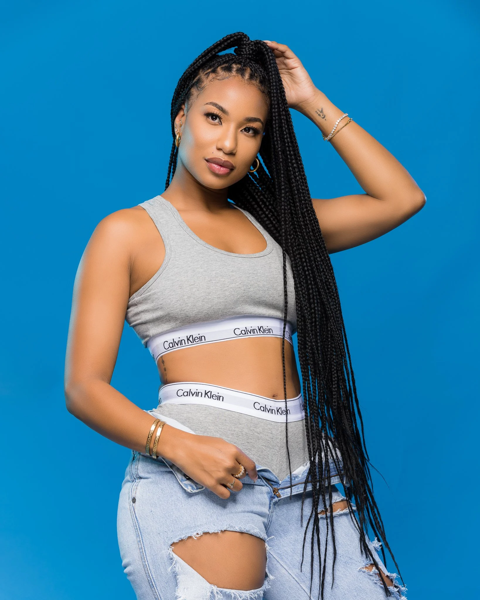 Young woman with long braids wearing a gray Calvin Klein sports bra and ripped denim shorts standing against a blue background.