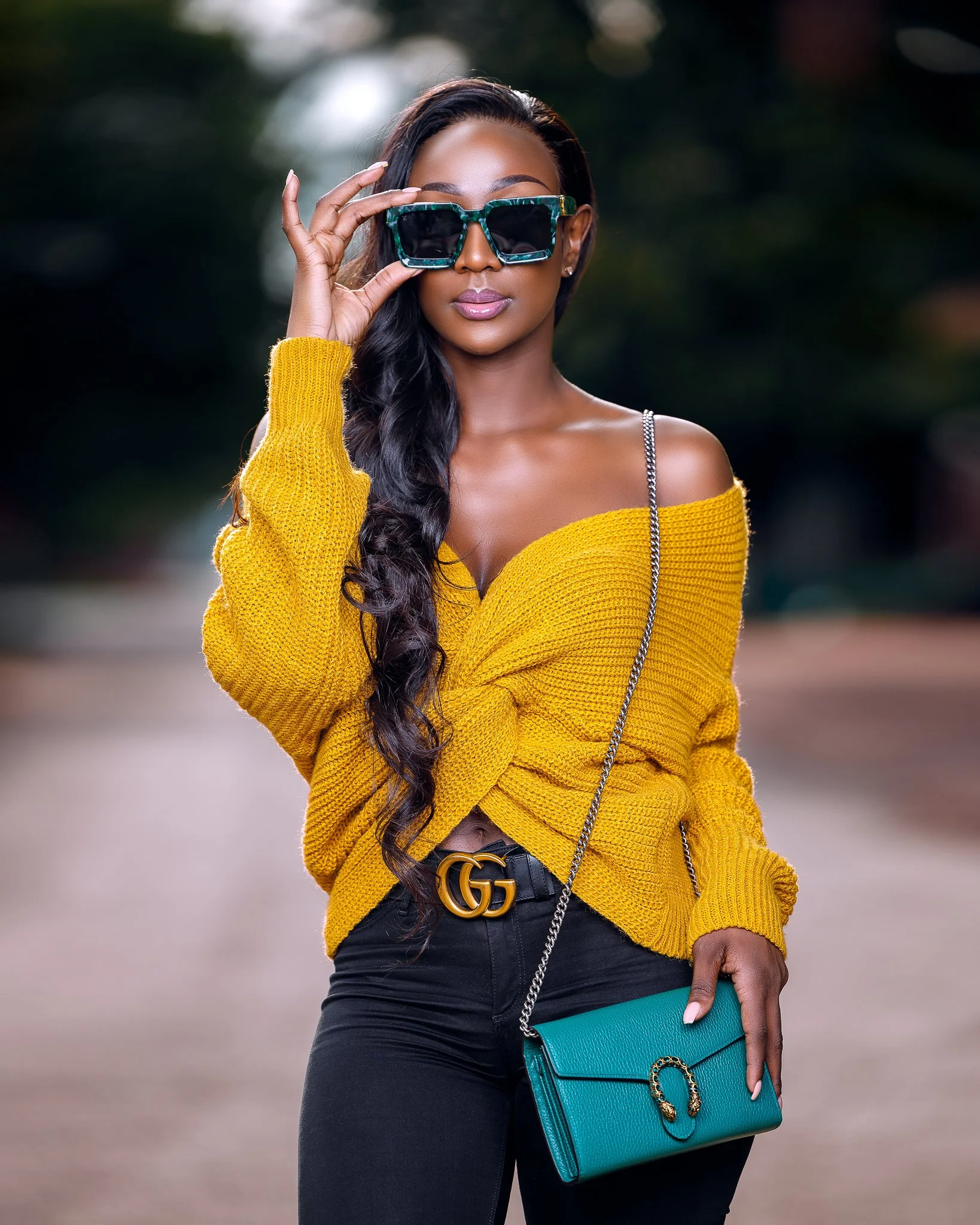 Woman wearing sunglasses and a yellow sweater, holding a turquoise clutch bag with a chain strap, standing outdoors.